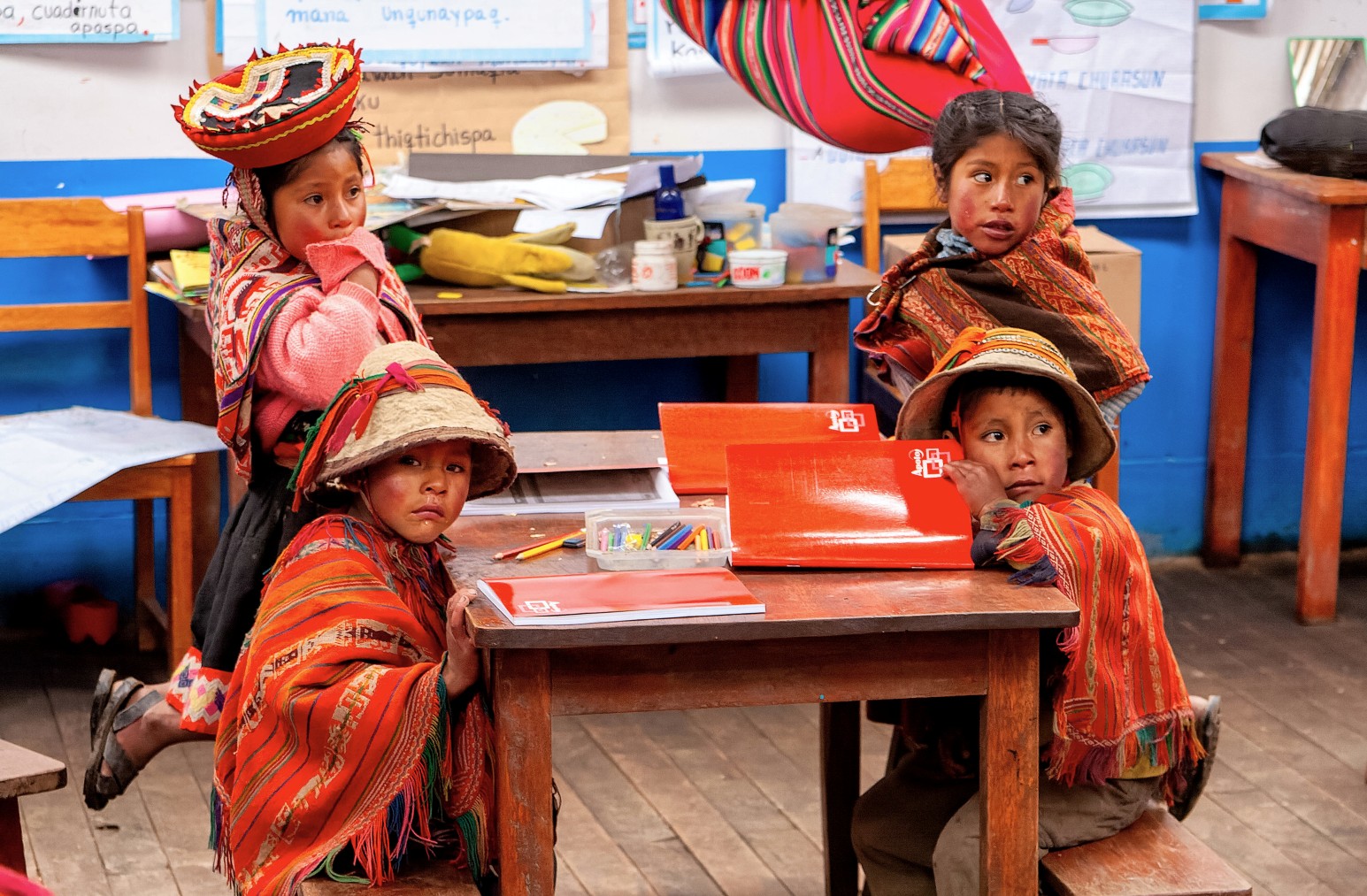Four children in traditional Andean dress sitting at a wooden desk in a classroom in Ollantaytambo