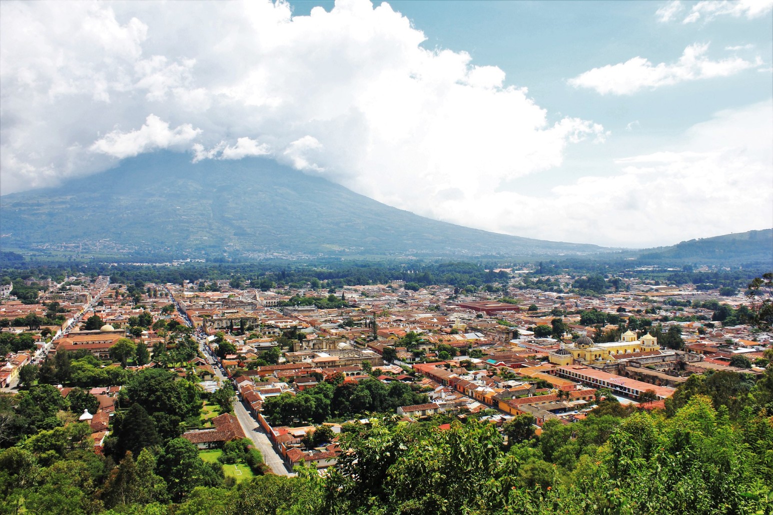 Aerial view over the terracotta rooftops and colonial churches of Antigua, Guatemala, with a large cloud-covered volcano towering over the city