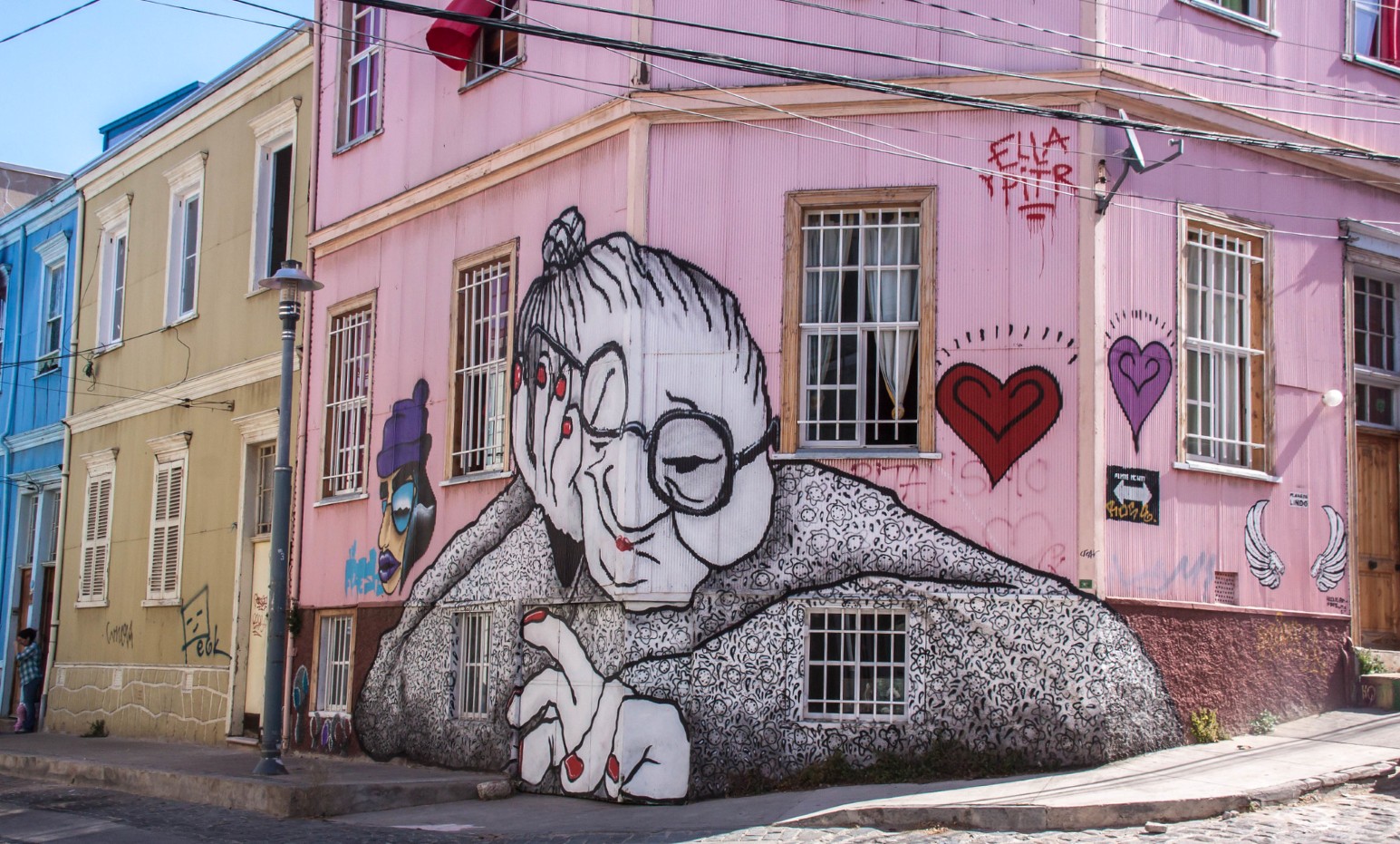 A large black and white mural of an elderly woman in glasses painted across the corner of a pink house in Valparaíso, with pastel blue and olive buildings running down the steep street beside it.