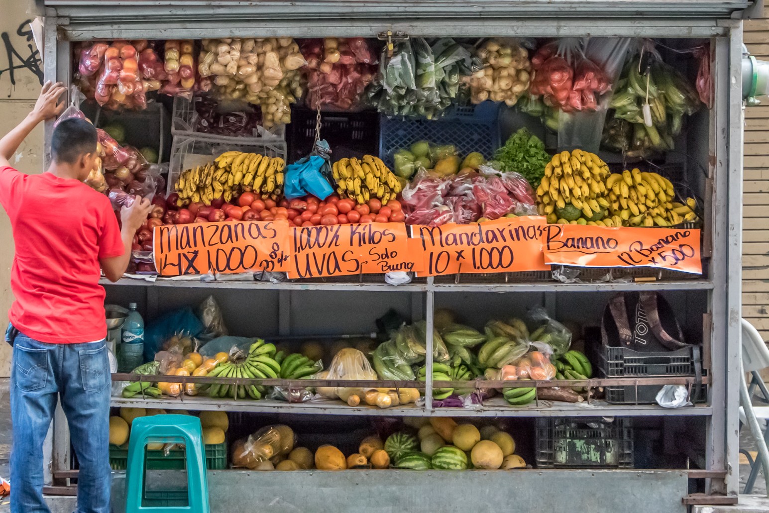 A vendor arranging produce at a street fruit stall stacked with bananas, tomatoes and apples, with handwritten price signs