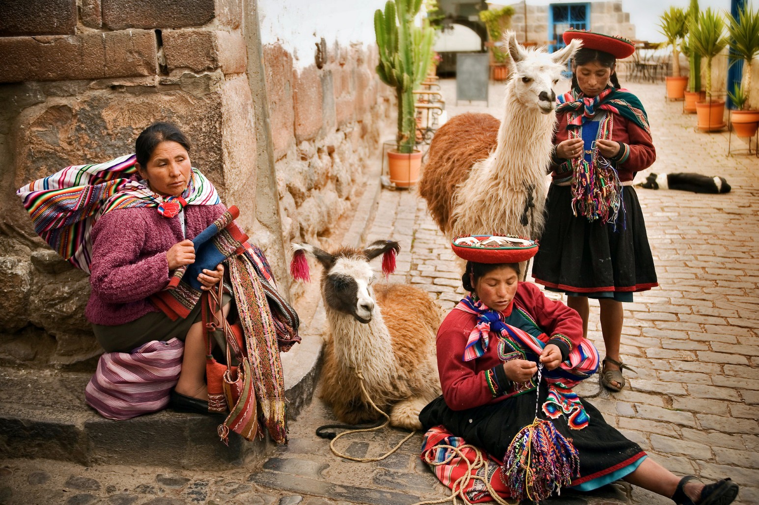 Two women in traditional Andean dress with llamas, sitting and standing on a cobblestone street