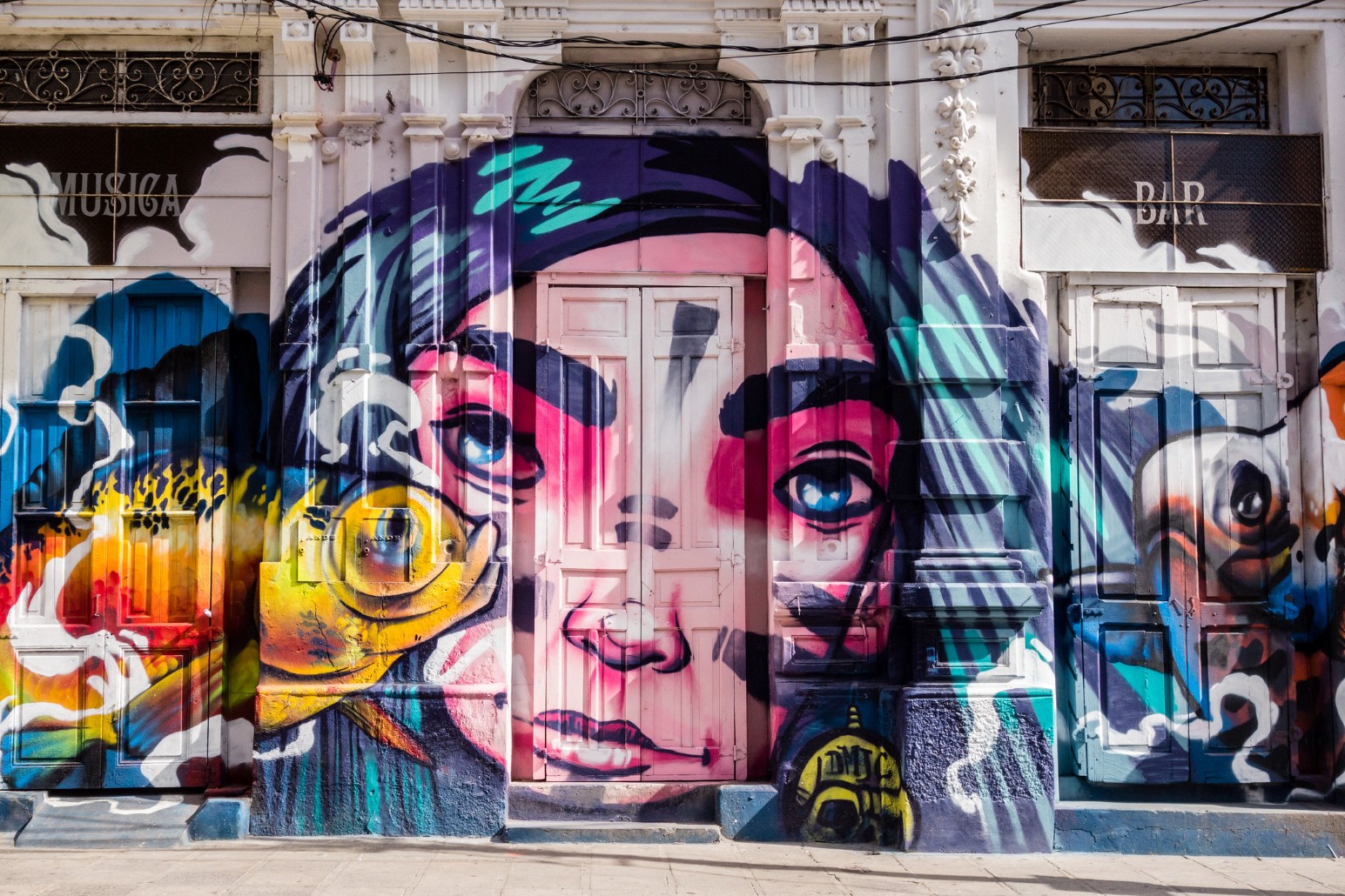 A large mural of a face in pink, purple and turquoise painted across the shuttered doorways of an old building in Asunción, between signs reading “Música” and “Bar”.