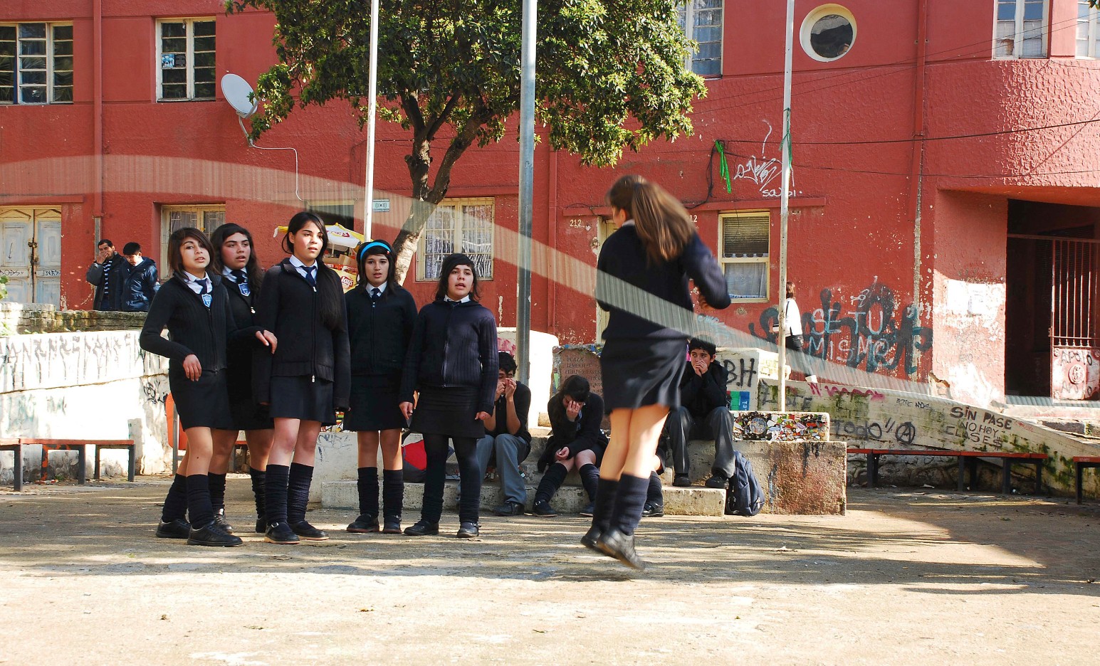 School pupils in dark uniforms gathered in a sunlit concrete yard in Valparaíso, one skipping in the foreground while others watch from steps in front of a red building.