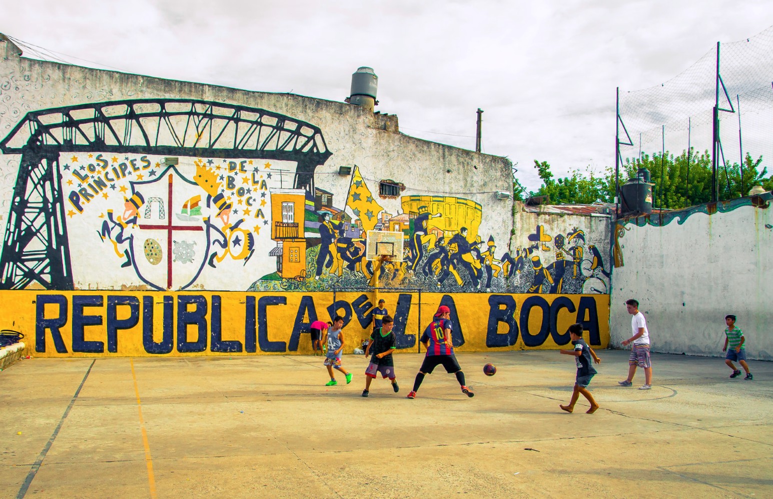 Children playing football on an outdoor court in La Boca, Buenos Aires, in front of a large blue and yellow mural reading “República de La Boca”