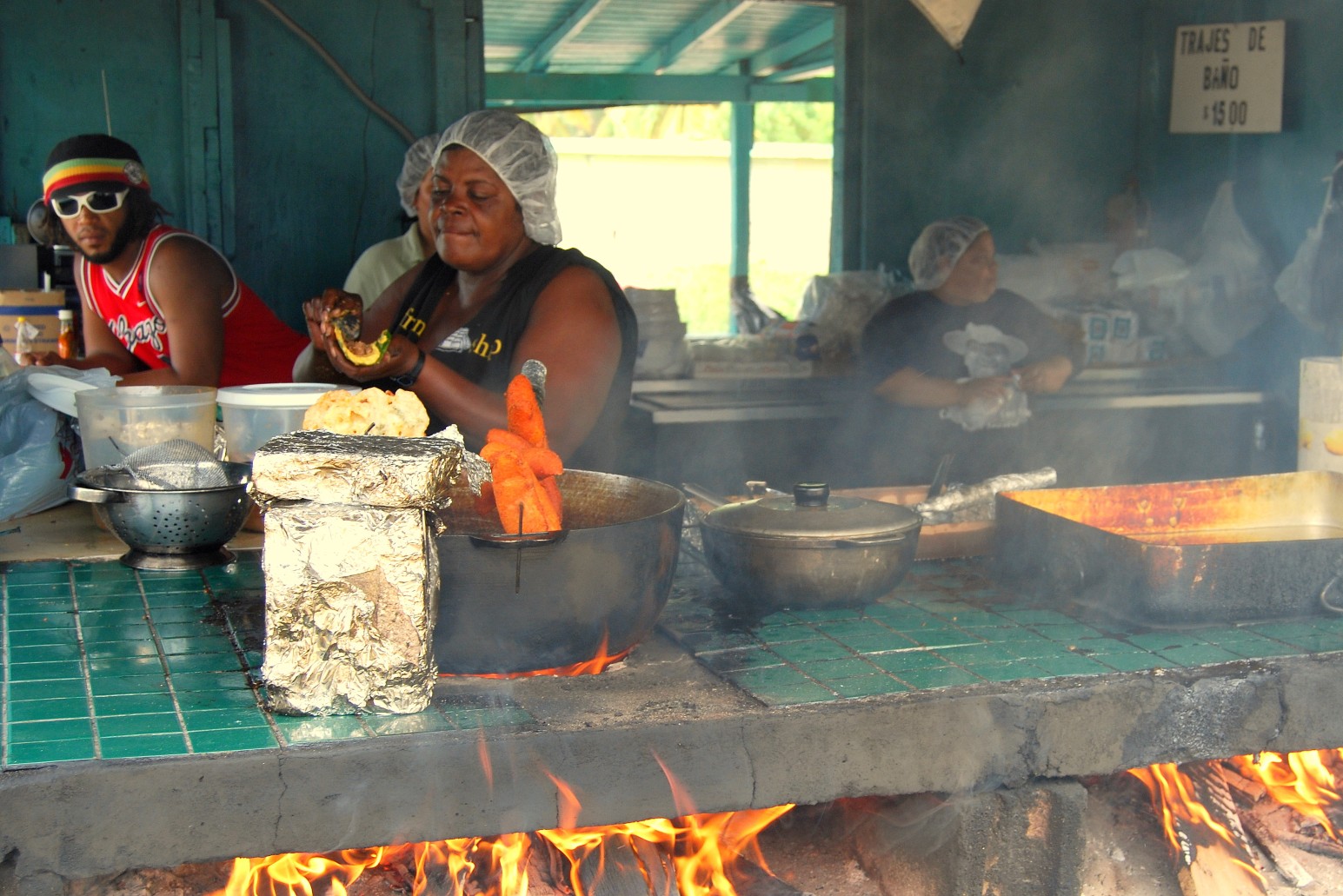 Cooks preparing food over an open wood fire at a roadside kiosk in Piñones, Puerto Rico, with frying pans, skewers and smoke rising over the counter.
