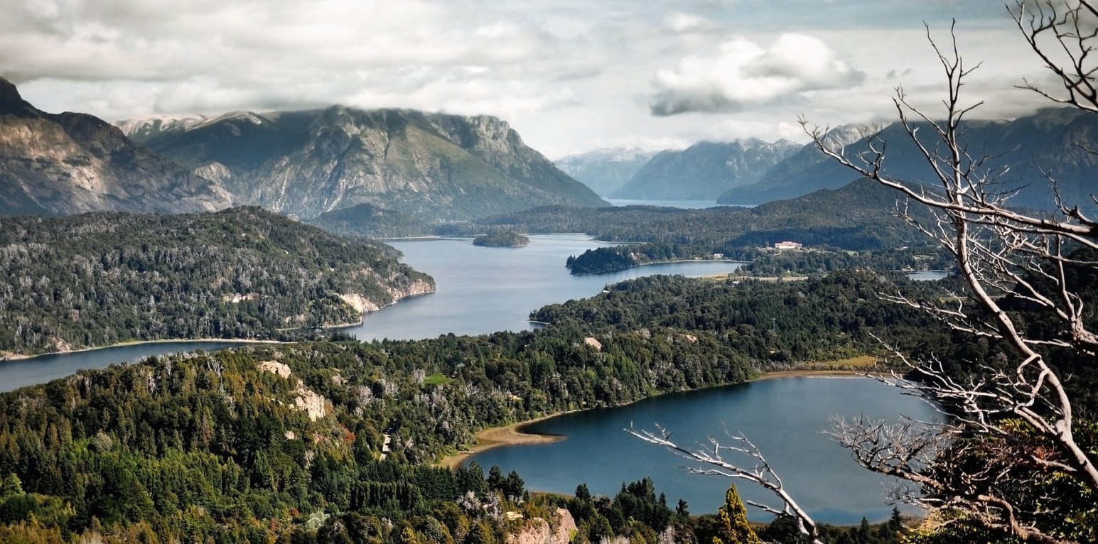 View over deep blue lakes and forested peninsulas near Bariloche, Argentina, with rugged Andes mountains under low clouds and bare branches in the foreground