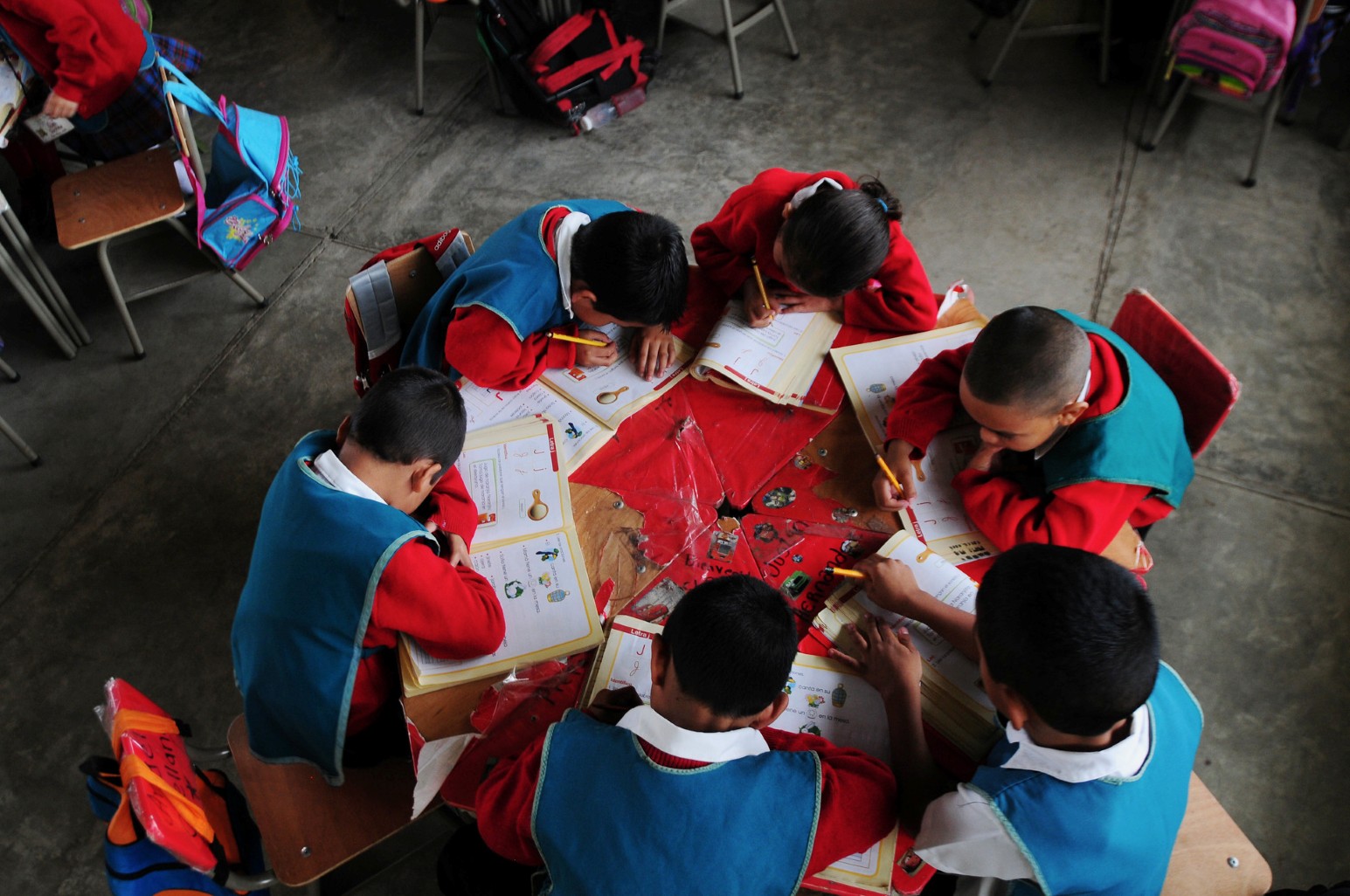Overhead view of young pupils in red and blue school uniforms working in their exercise books around a shared table at El Renacimiento School in Villa Nueva, Guatemala