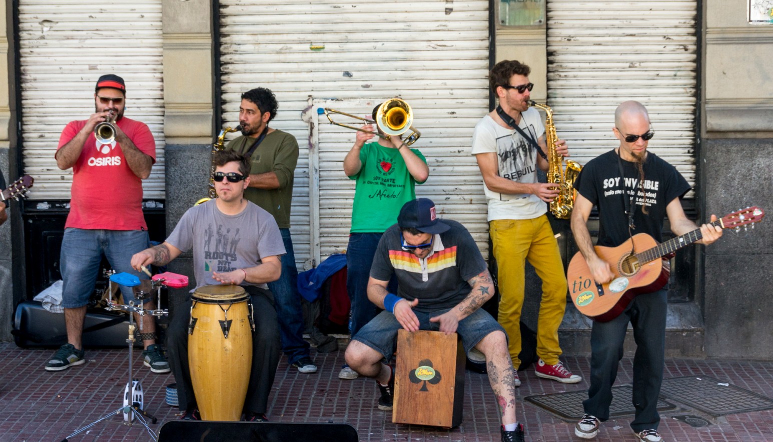 A street band performing on a pavement in Argentina, with musicians playing trumpet, saxophones, trombone, congas, cajón and acoustic guitar in front of closed shop shutters