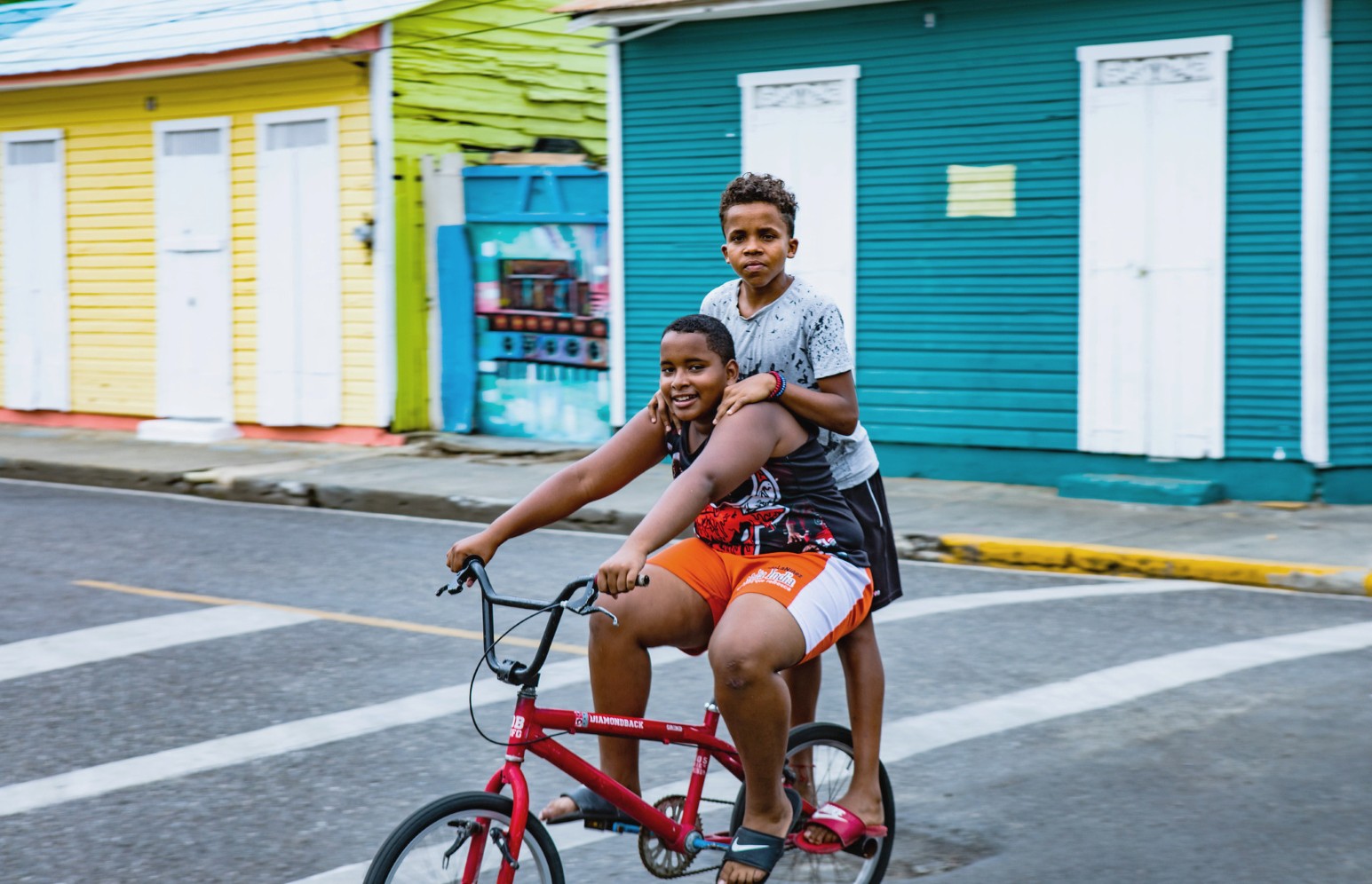 Two boys riding together on a red BMX bike along a street in Santiago, Dominican Republic, past brightly painted yellow, green and teal wooden houses