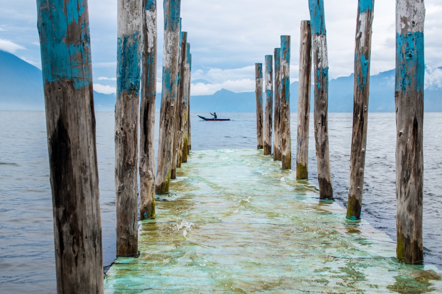 Weathered wooden posts painted turquoise lining a submerged jetty on Lake Atitlán, Guatemala, with a person paddling a small boat and volcanoes in the misty distance