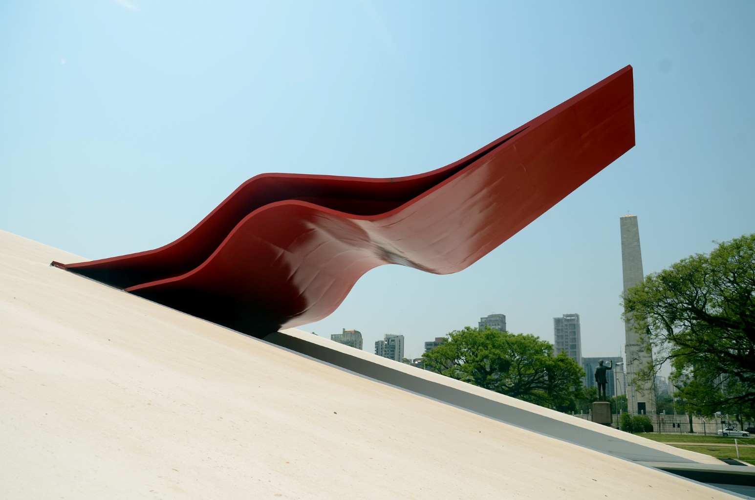 A red wave-shaped sculpture on a sloped monument in São Paulo, with the city skyline and an obelisk in the background
