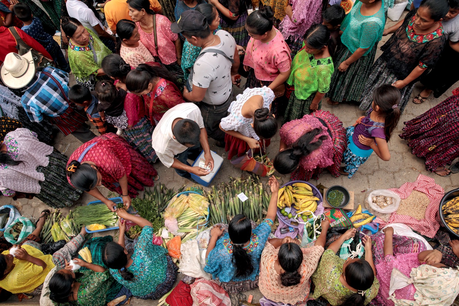 Overhead view of a busy market in Tucurú, Guatemala, with women in colourful traditional Mayan clothing selling bananas, spring onions and fresh produce laid out on the ground
