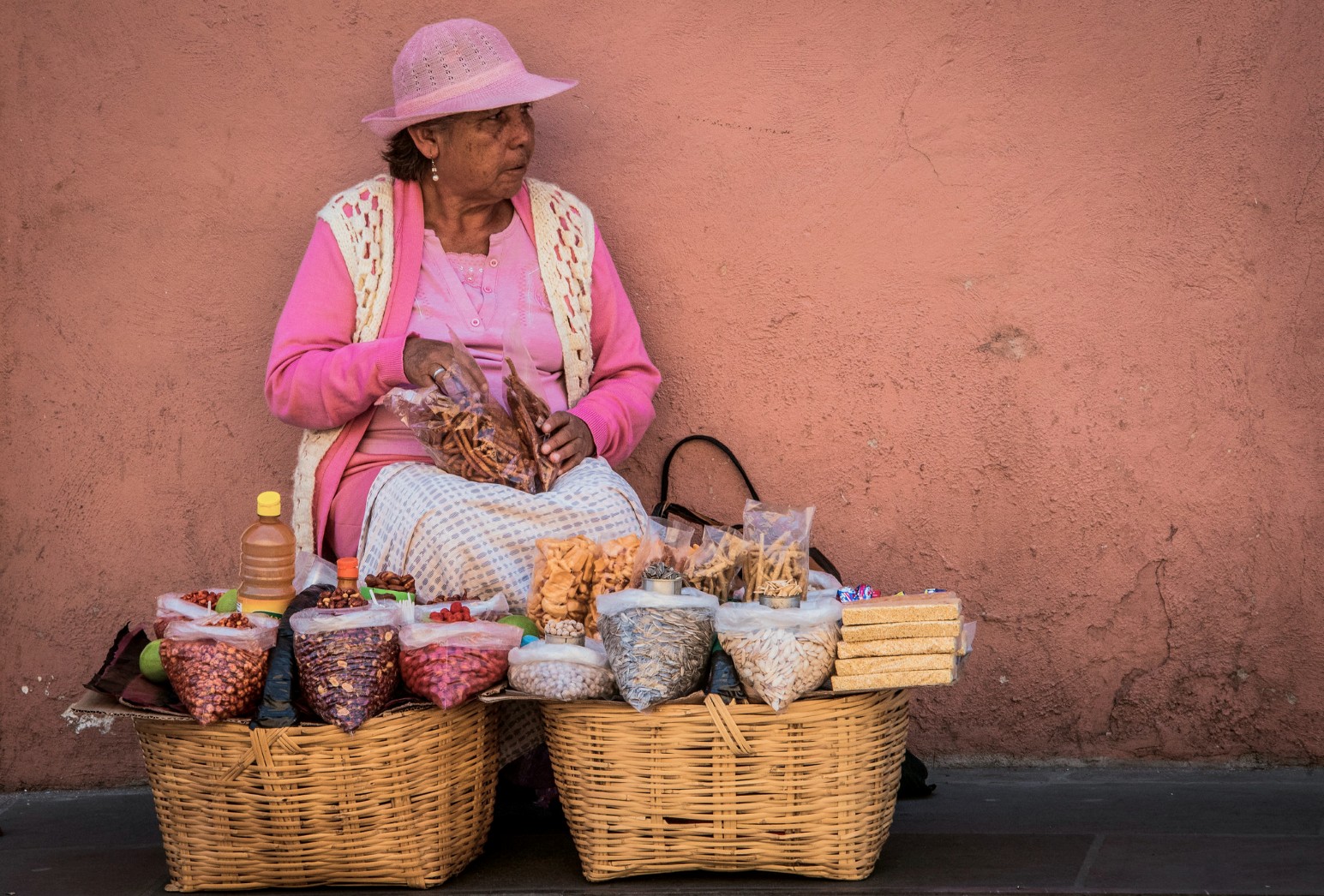 A street vendor dressed in pink sitting between two large baskets of nuts, seeds and snacks against a pink wall in Puebla