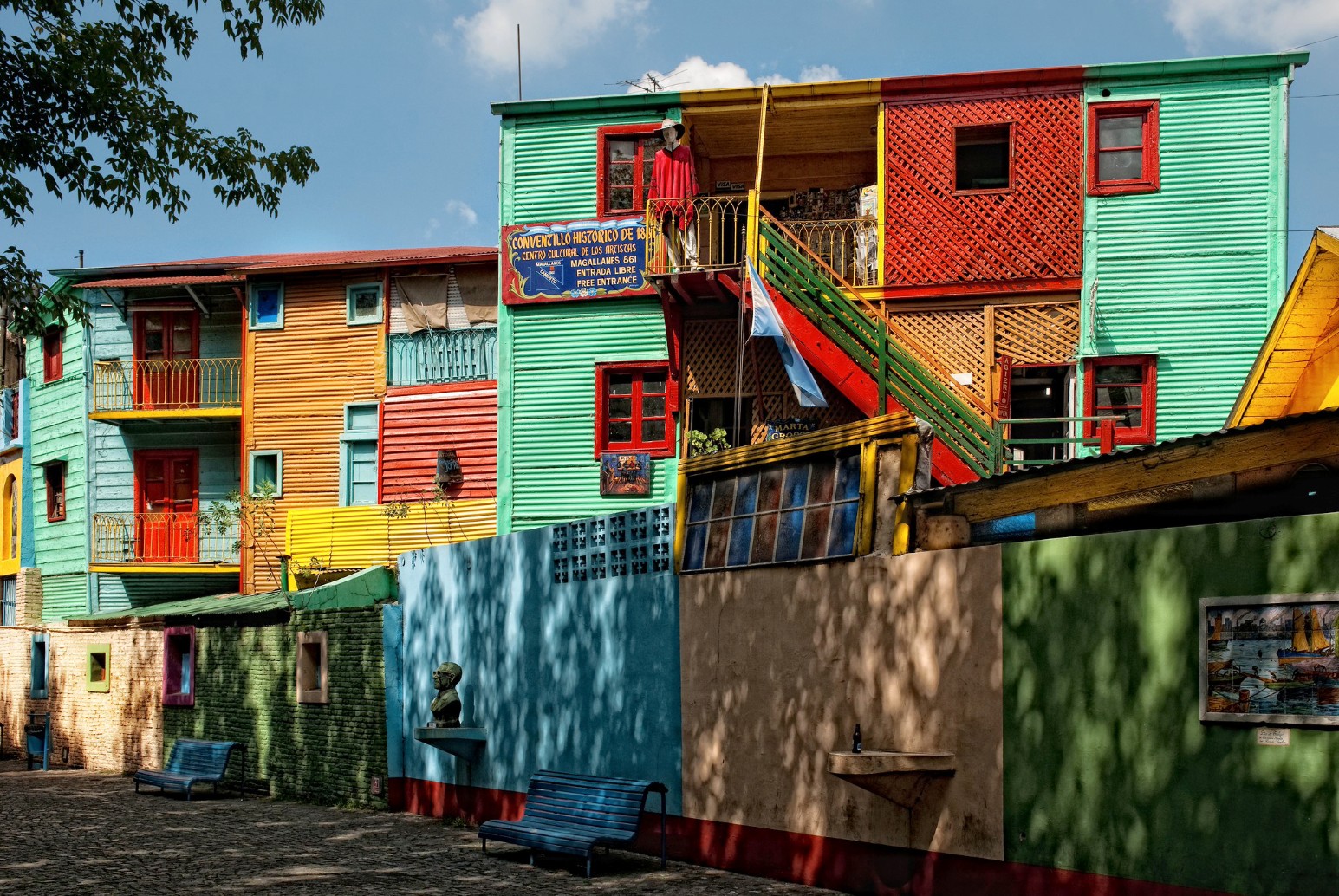 Brightly painted corrugated metal houses in green, red, orange and yellow in the Caminito area of La Boca, Buenos Aires, with an Argentine flag hanging from a balcony