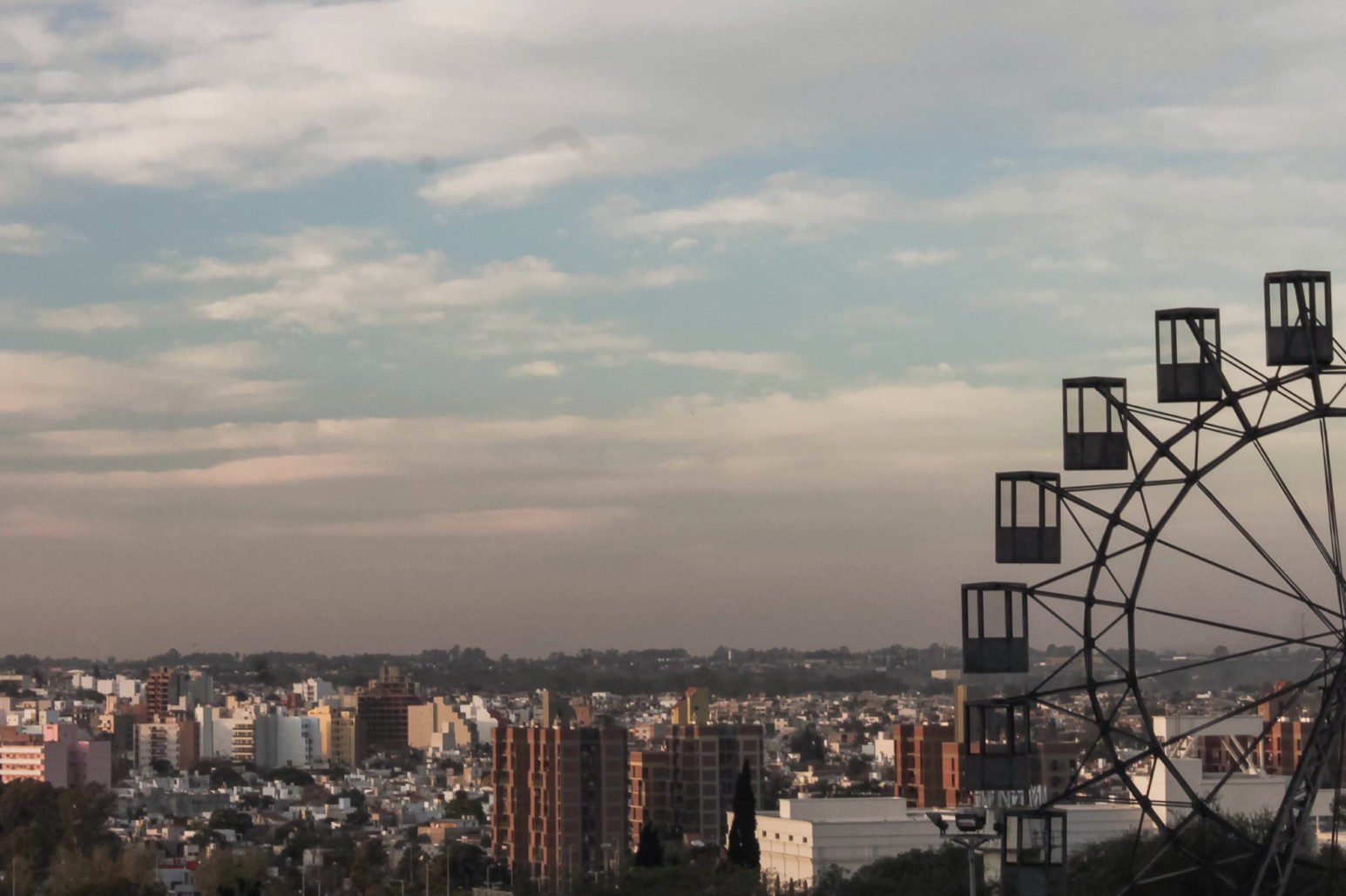 The skyline of Córdoba, Argentina, stretching to the horizon under a soft cloudy sky, with the silhouette of a Ferris wheel in the foreground