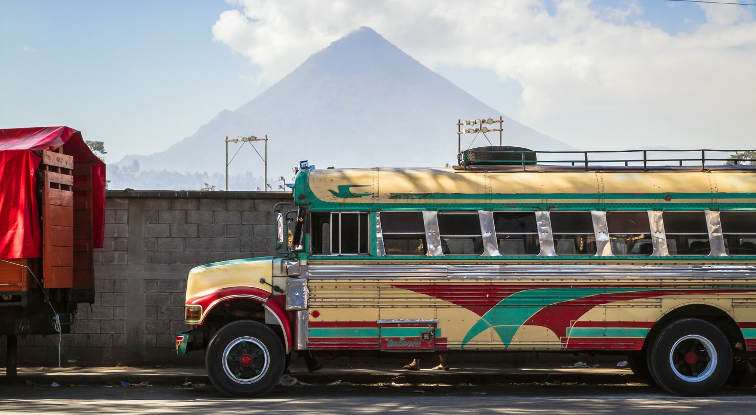 A brightly painted yellow, red and green chicken bus parked on a street in Quetzaltenango, Guatemala, with a cone-shaped volcano rising behind it