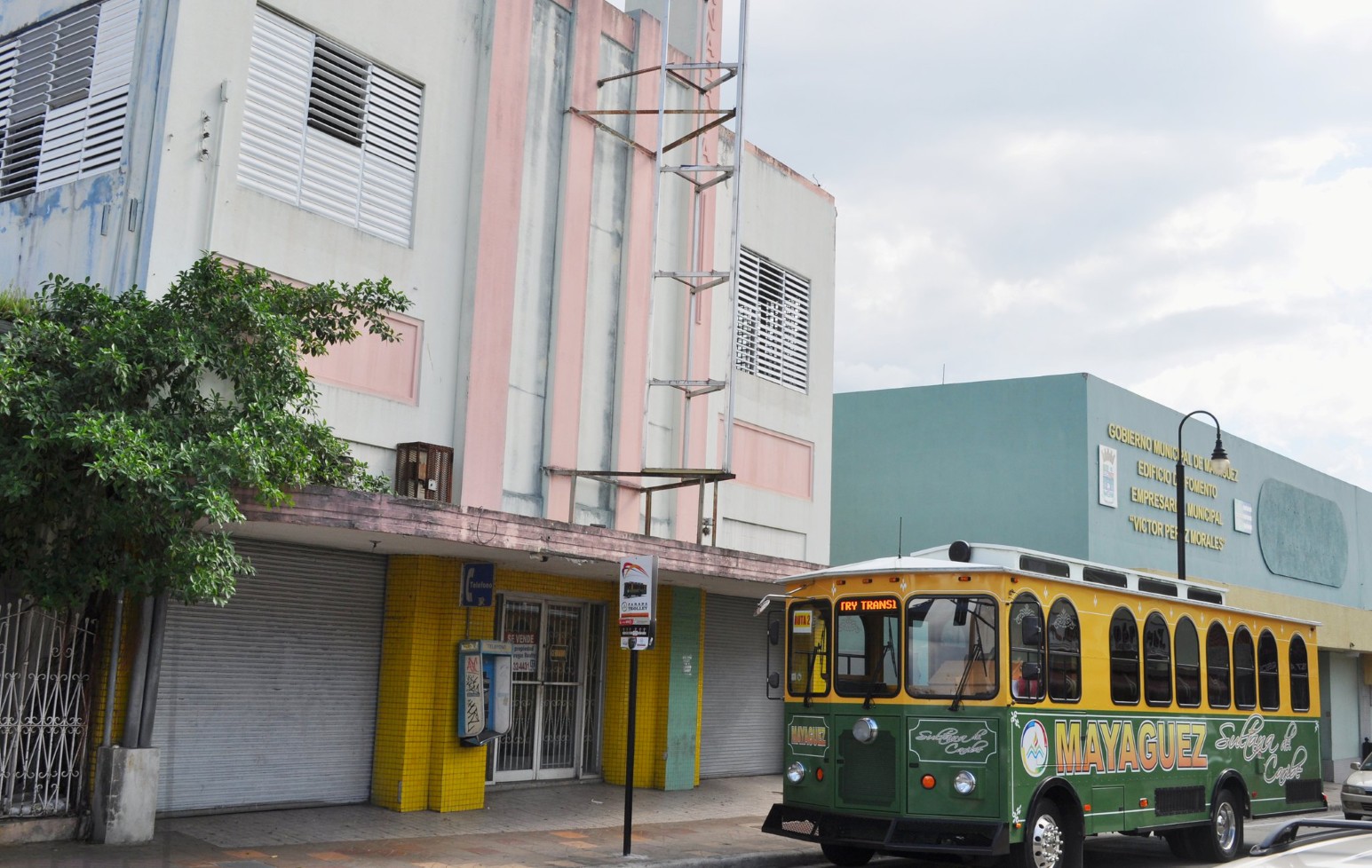 A green and yellow Mayagüez trolley bus parked outside pastel art deco buildings and the municipal government offices in downtown Mayagüez, Puerto Rico.