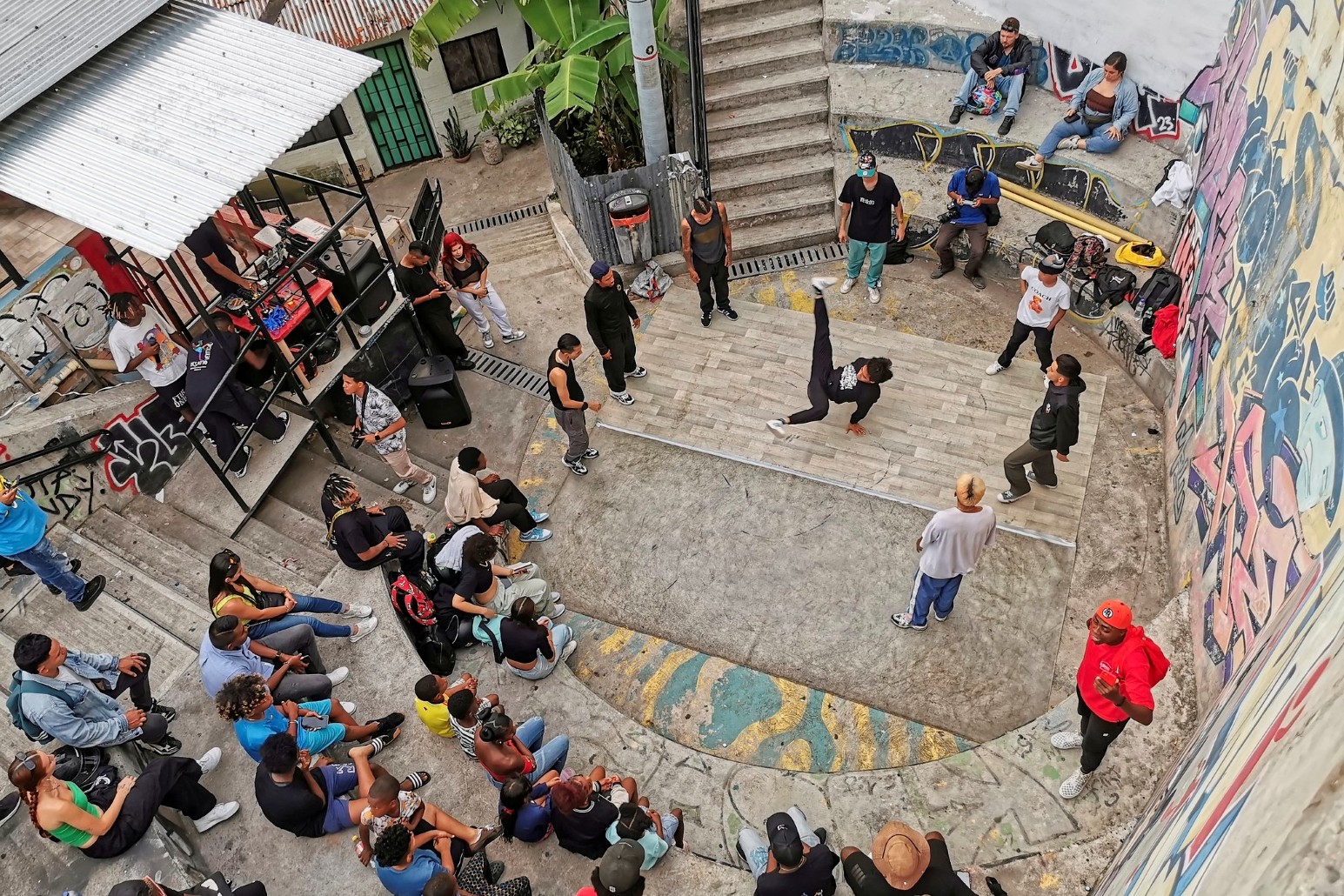 Breakdancers performing in a graffiti-covered courtyard in Medellín while a crowd watches from the steps