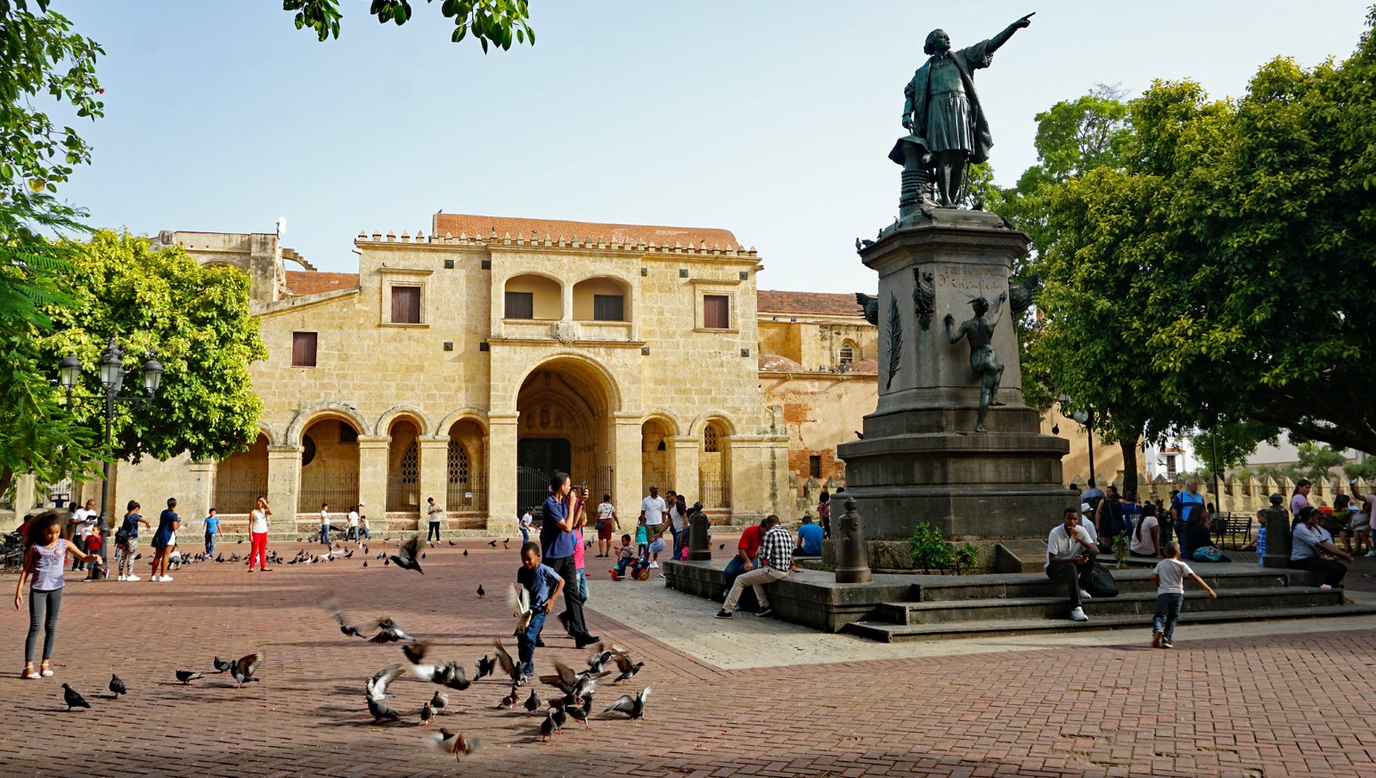 Families and children among pigeons in Parque Colón, Santo Domingo, with the statue of Christopher Columbus pointing out over the square in front of the stone Catedral Primada de América