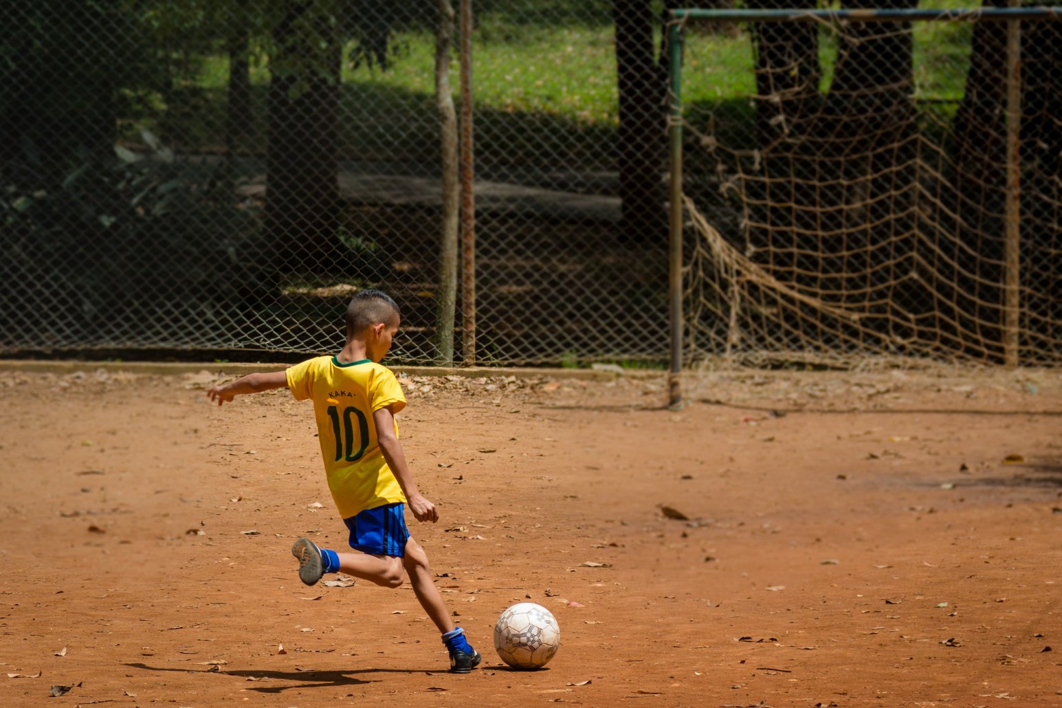 A young boy in a yellow football shirt kicking a ball on a dirt pitch