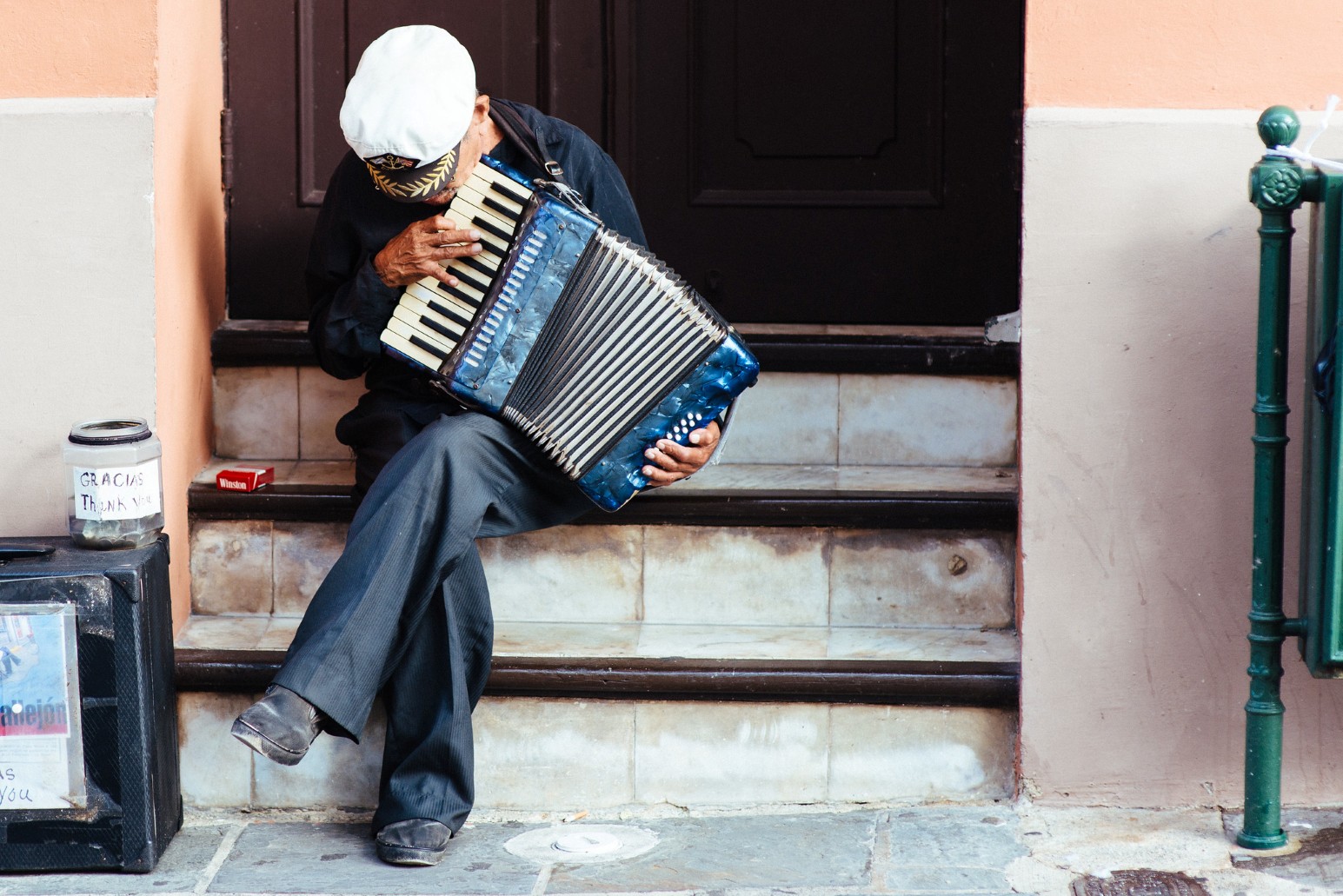 An elderly street musician playing a blue accordion on a doorstep in Old San Juan, with a tip jar labelled “Gracias / Thank you” beside him.