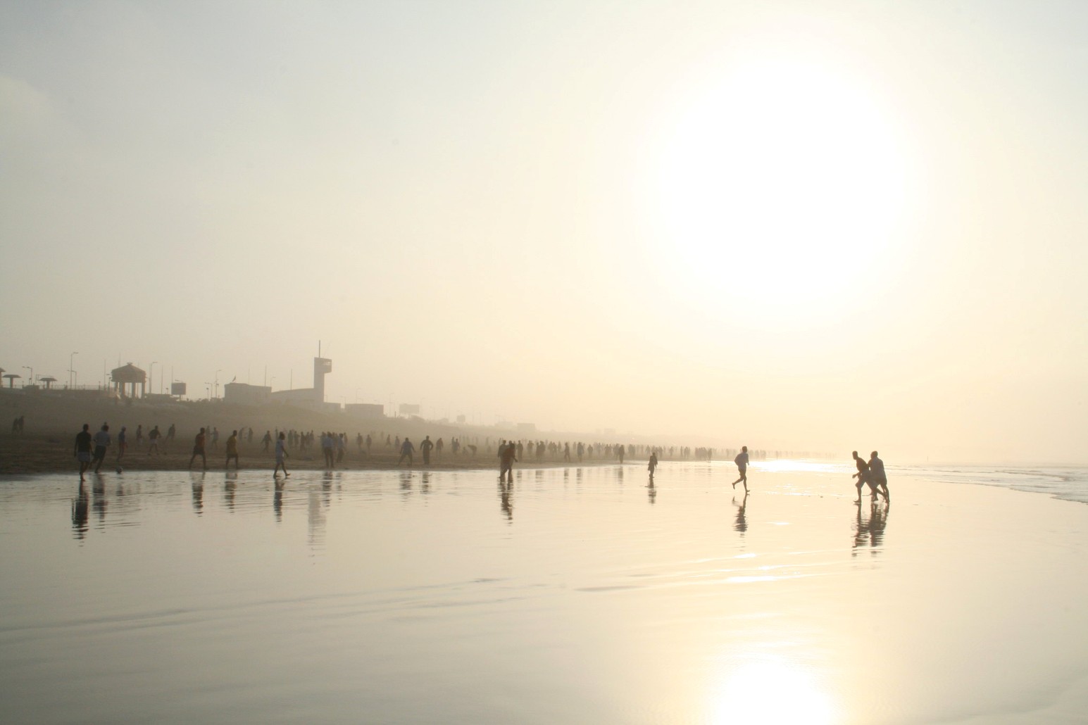 Silhouetted groups of people playing football on the wet sand of a hazy beach in Casablanca, Morocco, with their reflections on the shoreline and a low sun glowing through the mist