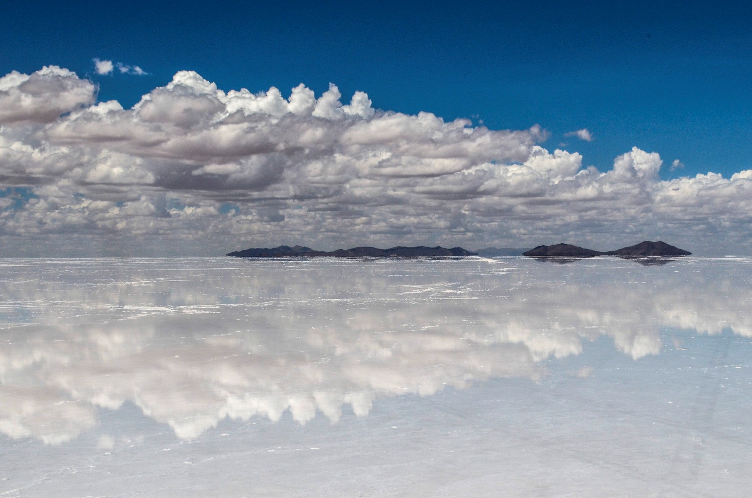 The Salar de Uyuni salt flat in Bolivia covered in a thin layer of water, creating a mirror-like reflection of white clouds and a deep blue sky, with low dark islands on the horizon