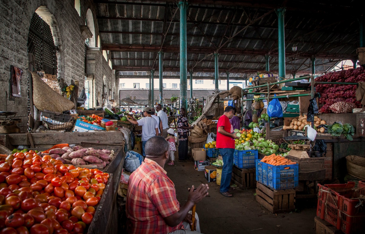 Shoppers and stallholders in a covered fruit and vegetable market in Mombasa, Kenya, with piles of tomatoes, sweet potatoes, carrots and pineapples beneath a high metal roof