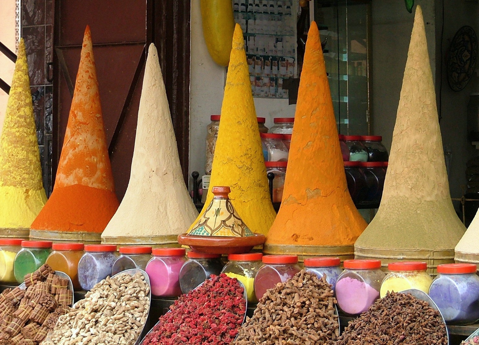 Tall cones of yellow, orange and beige ground spices on a market stall in Marrakesh, Morocco, with a decorated tagine, jars of coloured powders and scoops of dried spices in front