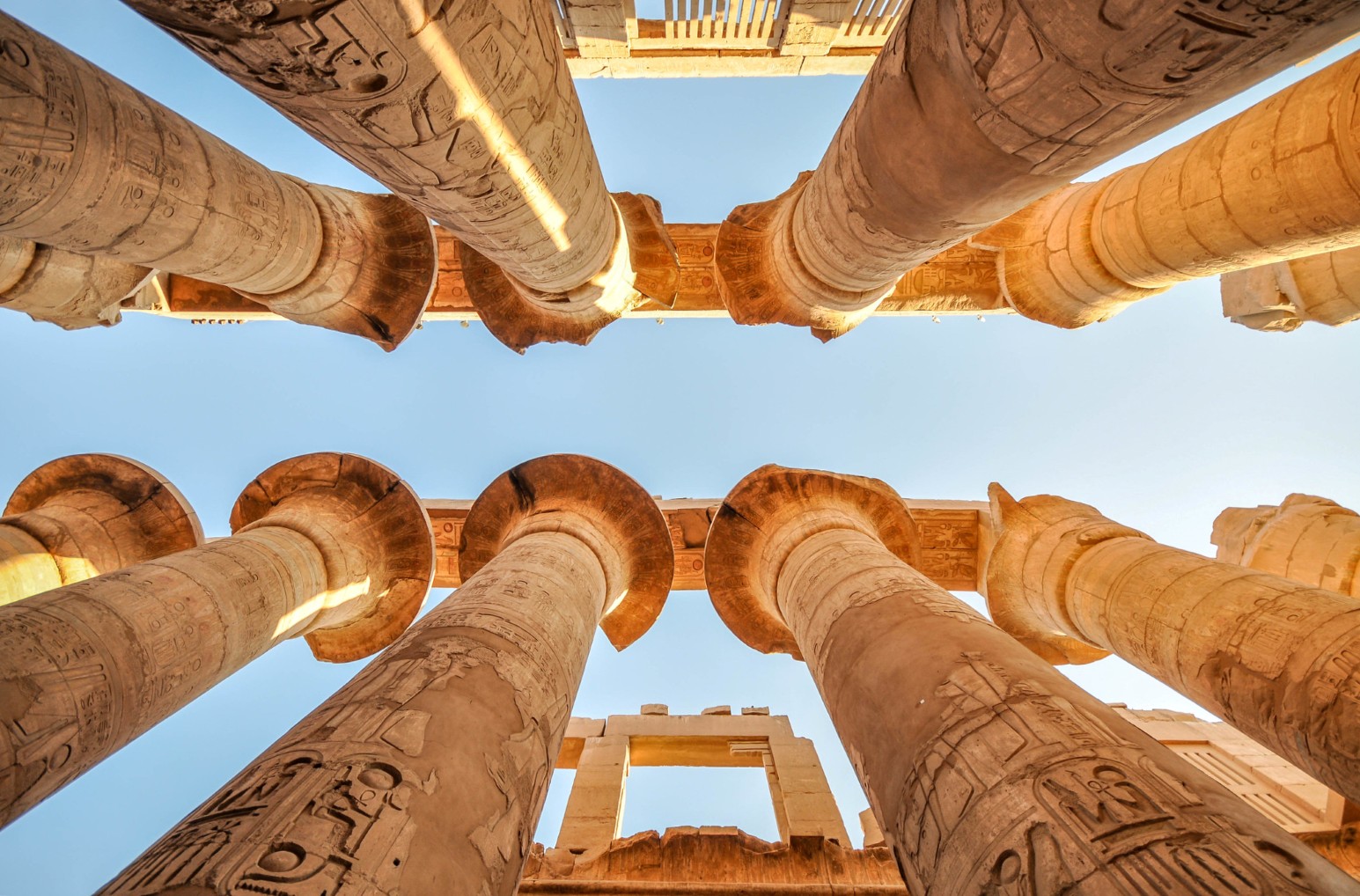 Looking up at giant carved papyrus columns against a blue sky in the Great Hypostyle Hall at Karnak Temple, Luxor, Egypt