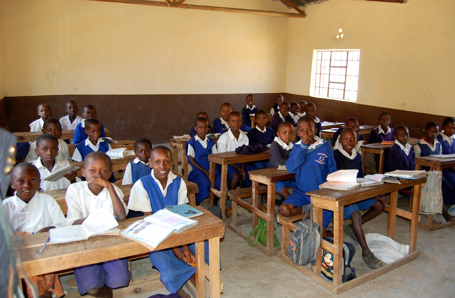 Smiling schoolchildren in blue and white uniforms sitting at wooden desks with open exercise books in a classroom in Kiima Kimwe, Kenya