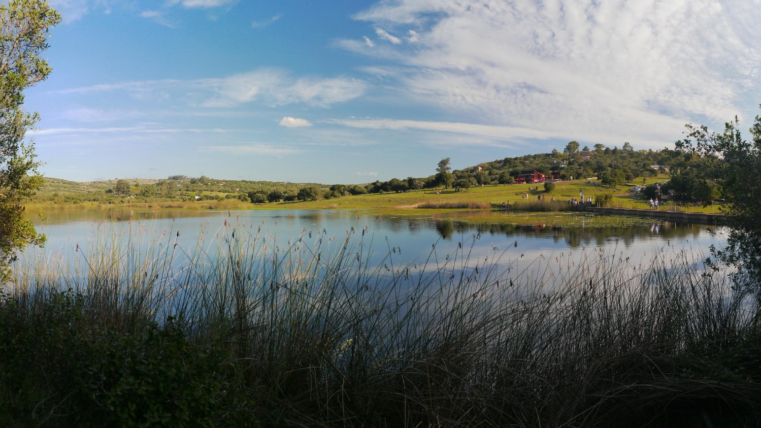 A calm lake in Villa Serrana, Uruguay, reflecting a blue sky, with tall reeds in the foreground and green wooded hills, a red-roofed building and people strolling along the far shore