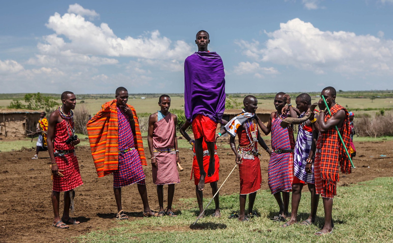 A Maasai man in a purple shuka leaps high in the air during a traditional jumping dance, watched by men in red and checked shukas in the Maasai Mara, Kenya