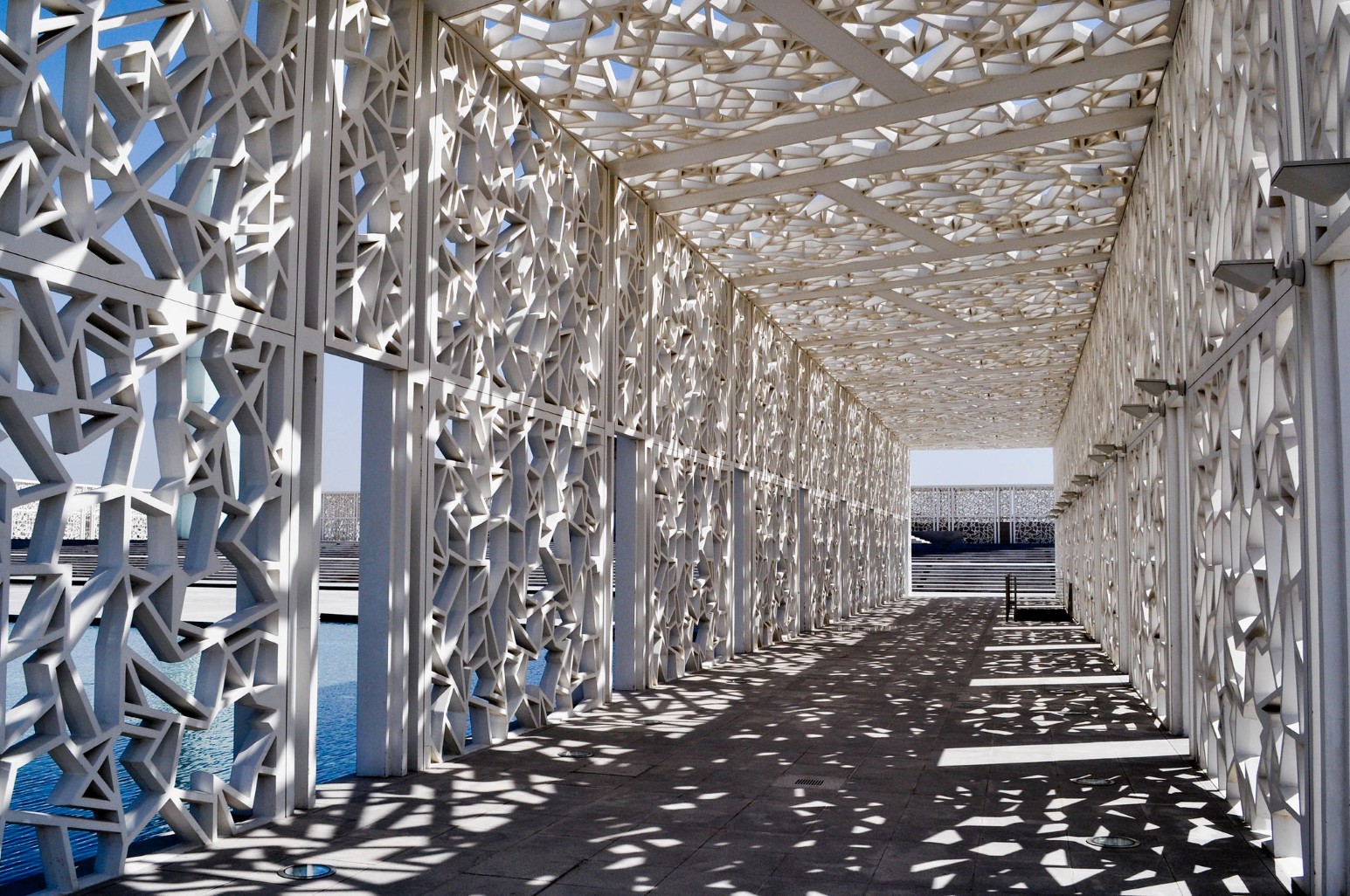 A covered walkway in Education City, Al Rayyan, Qatar, enclosed by white geometric lattice screens that cast intricate patterns of light and shadow across the floor
