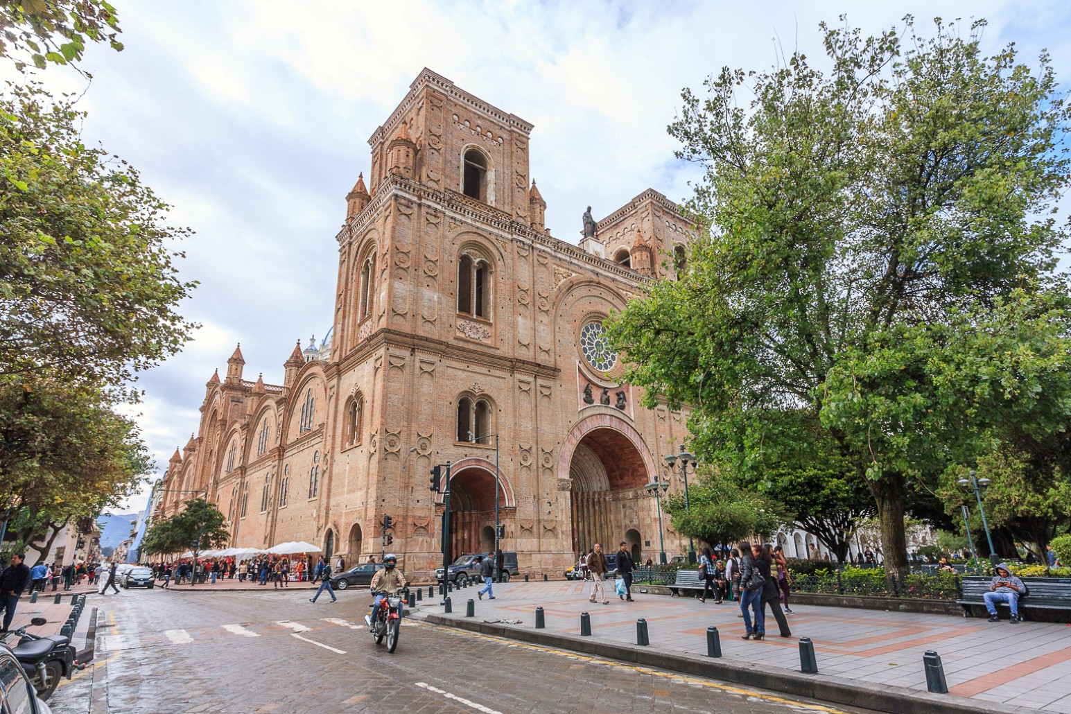 The red-brick New Cathedral of Cuenca, Ecuador, with its twin towers, rose window and arched entrances, seen from a tree-lined square with pedestrians and a passing motorcyclist