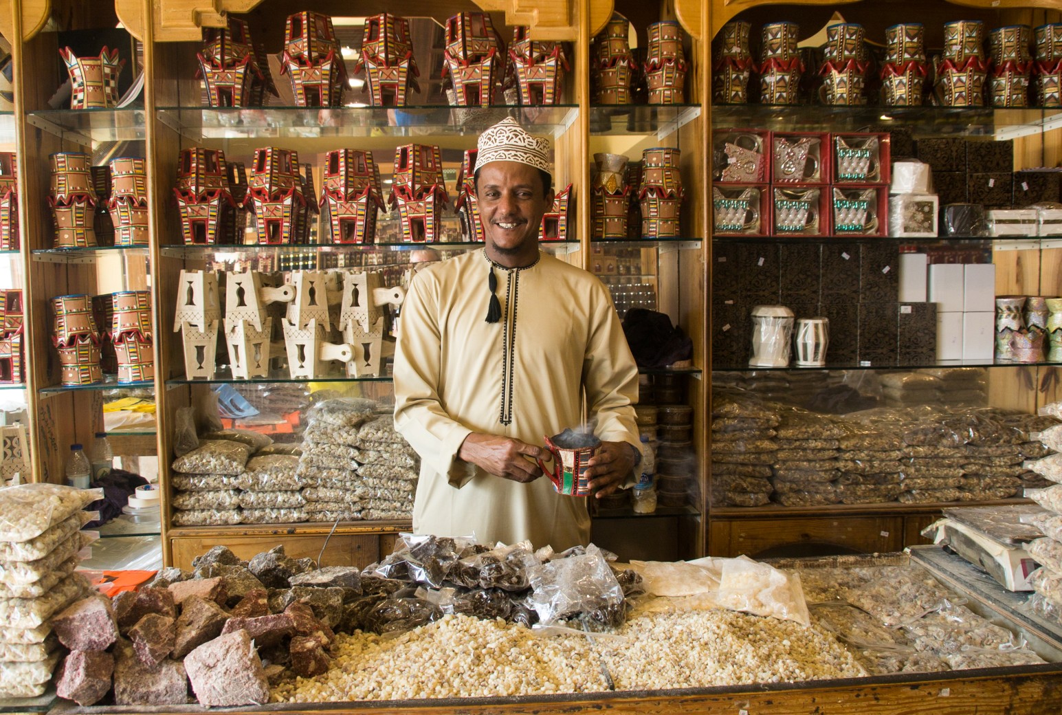 A smiling shopkeeper in a dishdasha and embroidered cap holds a smoking incense burner behind a counter piled with frankincense resin in Salalah, Oman, with shelves of painted burners behind him