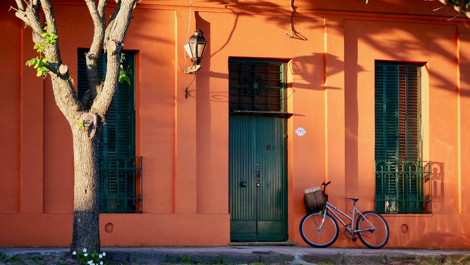 A bright orange colonial-style house in Colonia del Sacramento, Uruguay, with green shuttered windows, a green wooden door, a wall lantern and a bicycle with a basket leaning against the wall