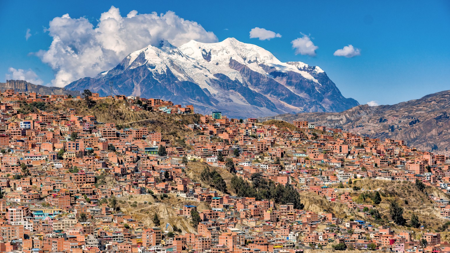 Red-brick houses packed across the hillsides of La Paz, Bolivia, beneath the snow-capped peaks of Mount Illimani and a bright blue sky