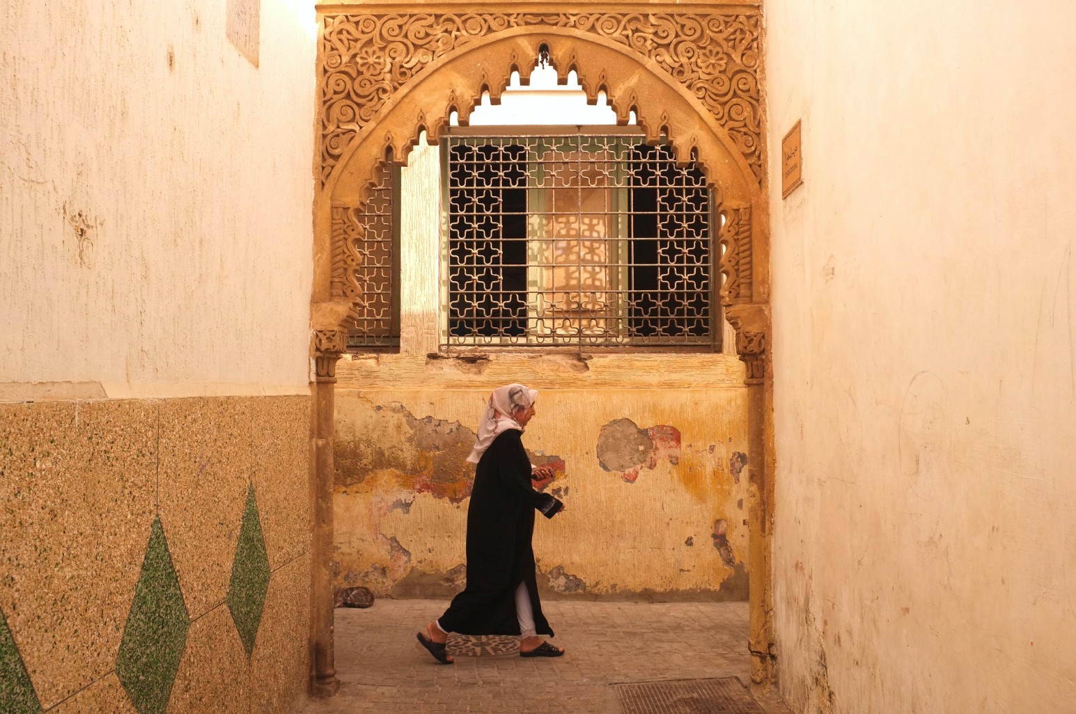 A woman in a black djellaba and white headscarf walks through a narrow lane in Rabat, Morocco, beneath an ornately carved stone archway framing a latticed window