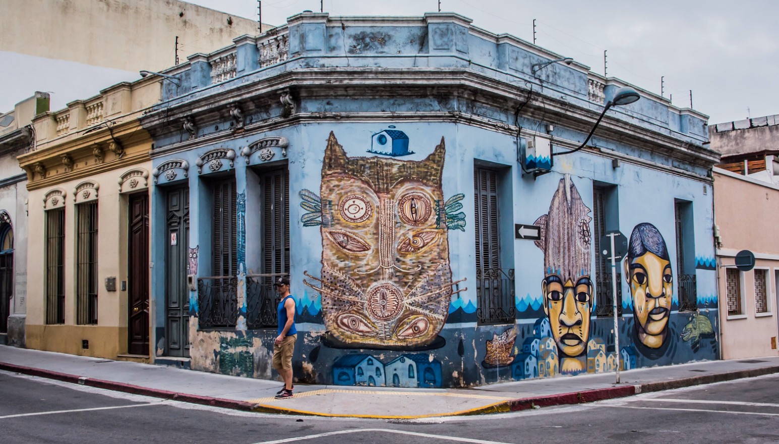 A man walks past a pale blue corner building in Montevideo, Uruguay, painted with colourful street art murals of a stylised cat face and two human faces