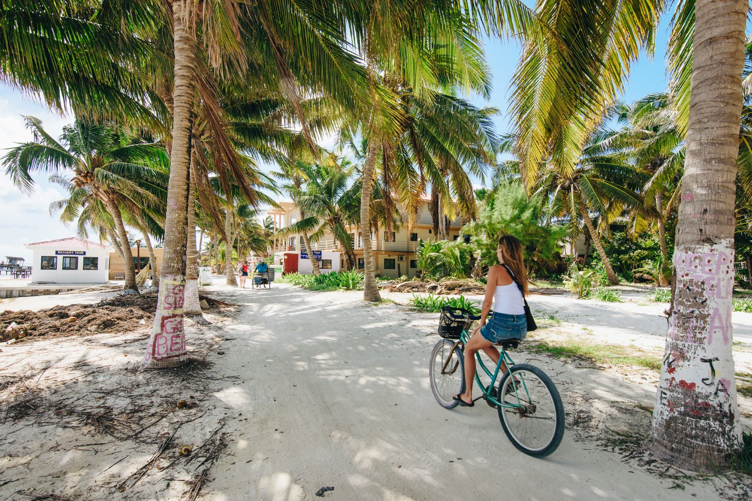 A woman cycling along a sandy, palm-shaded path on Caye Caulker, Belize, past low beachfront buildings and a tour booth