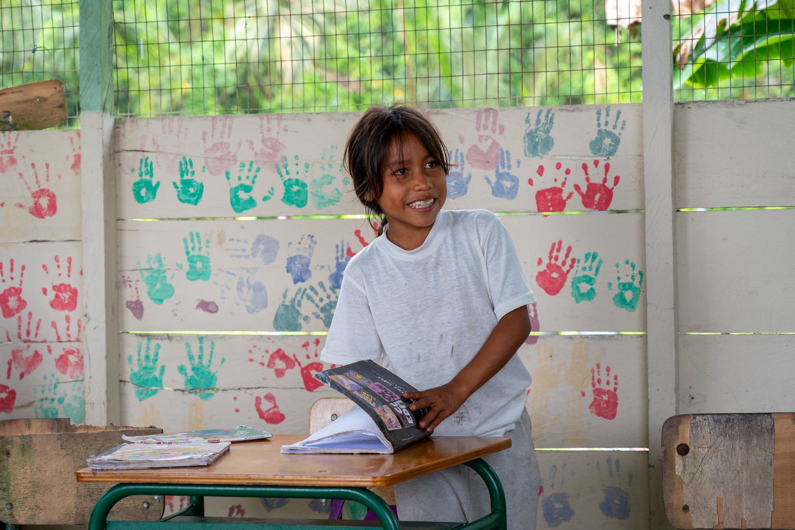 A smiling young girl standing at a wooden school desk with open exercise books in a classroom in Ecuador, in front of a wall covered in colourful painted handprints