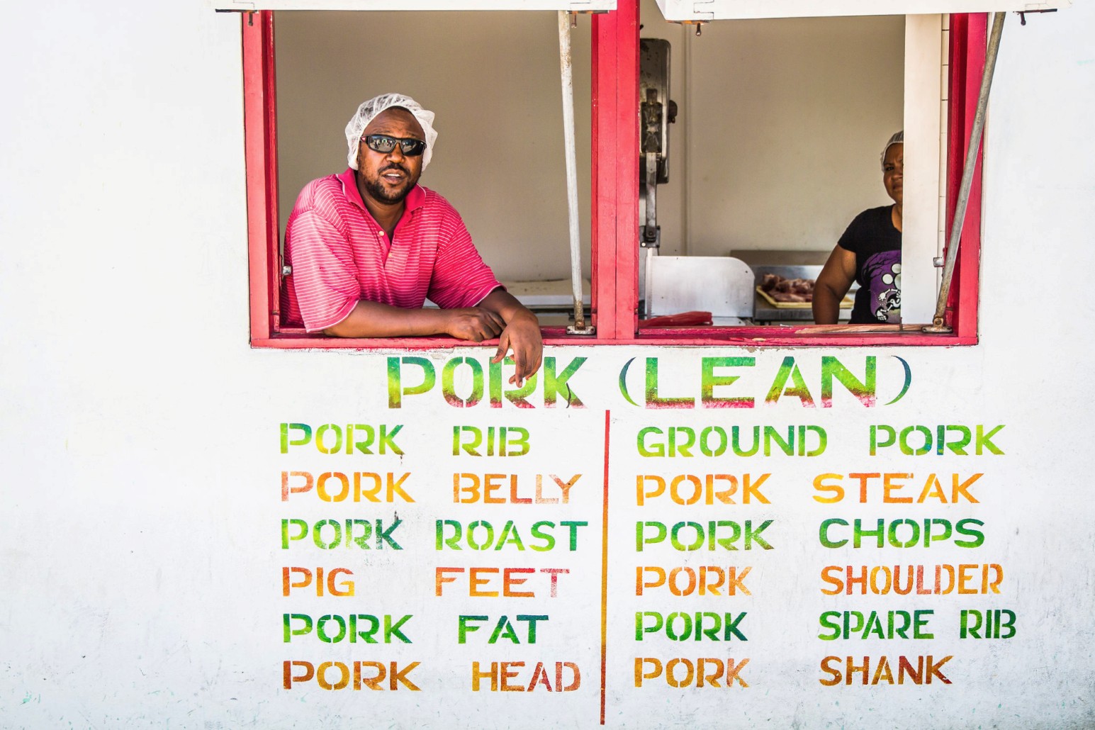 A butcher leaning out of the red-framed window of a meat stall in Belize City, above a brightly stencilled menu of pork cuts such as ribs, belly, chops and shank