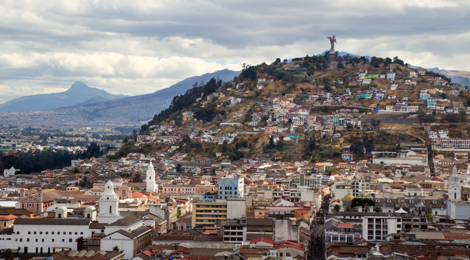 View across the rooftops and white church towers of Quito’s historic centre, Ecuador, towards El Panecillo hill and its winged Virgin statue, with Andean mountains in the background