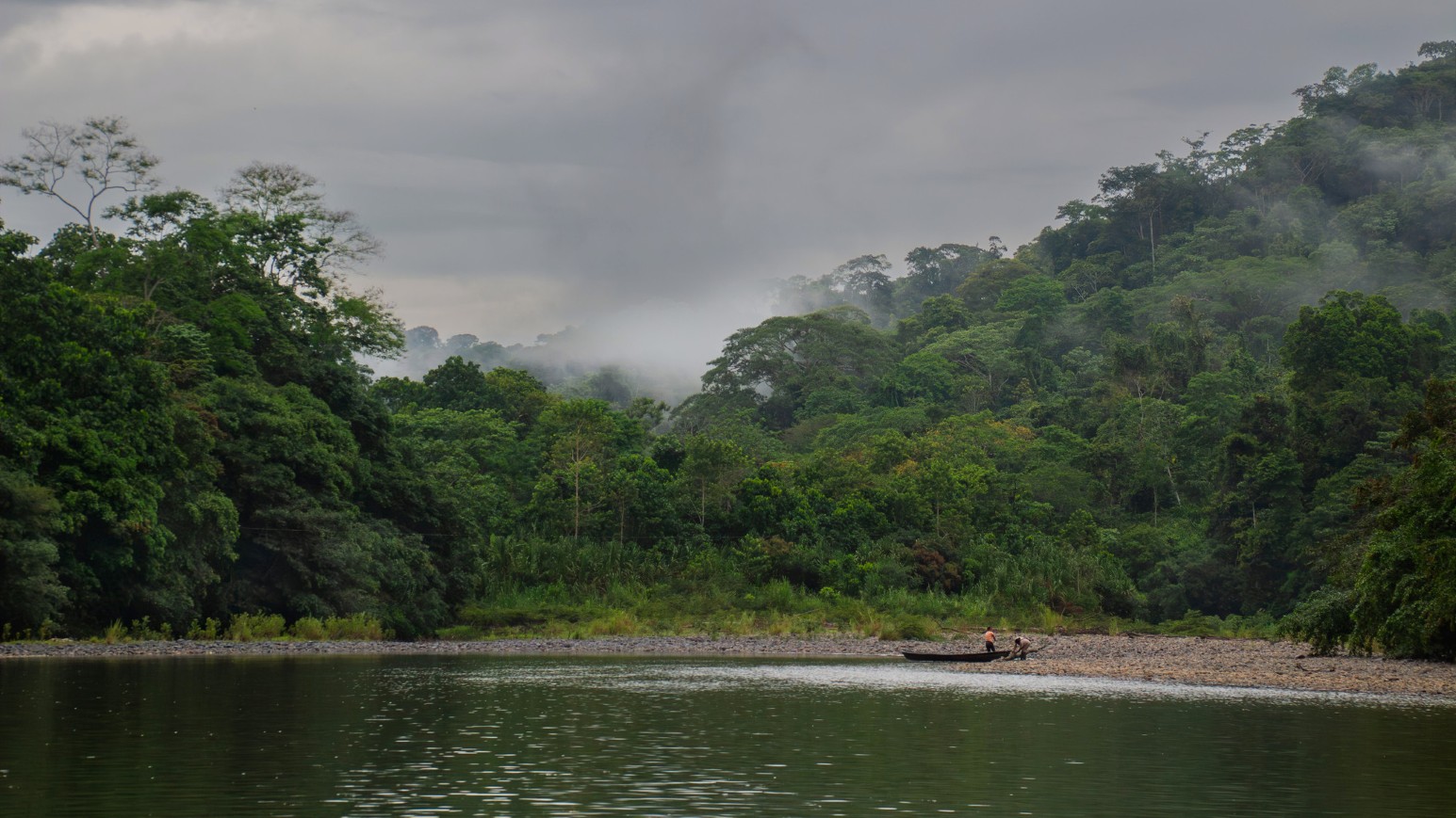 Two people beside a wooden canoe on a pebbly riverbank, beneath dense, mist-covered rainforest under a grey sky in Ecuador