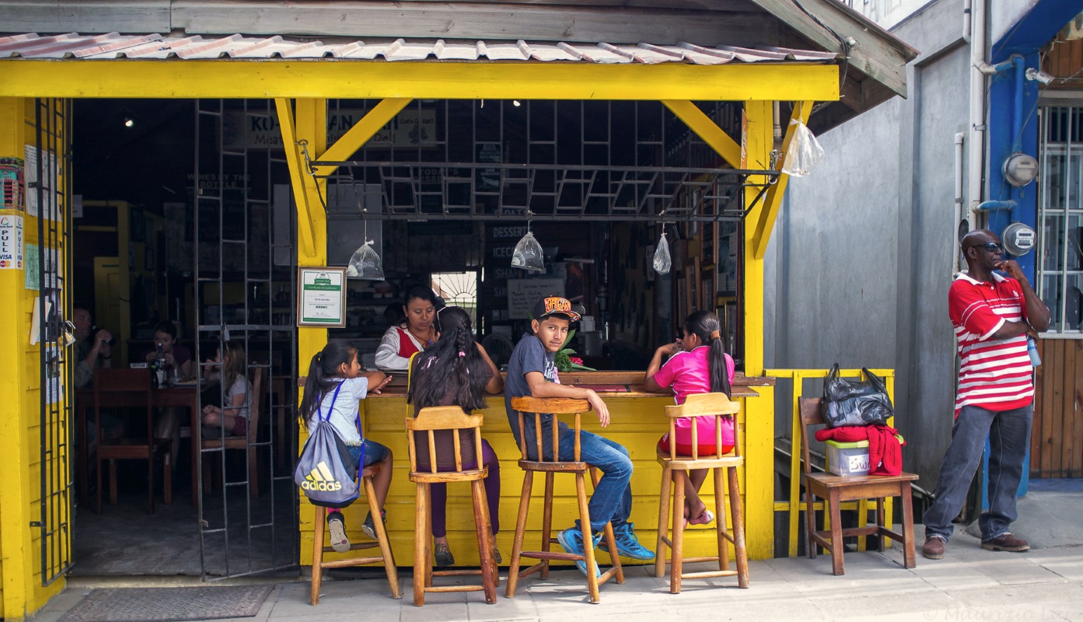 Children sitting on wooden stools at the counter of a bright yellow wooden café in Belize, with a woman serving behind the counter and a man in a red striped shirt standing on the pavement nearby