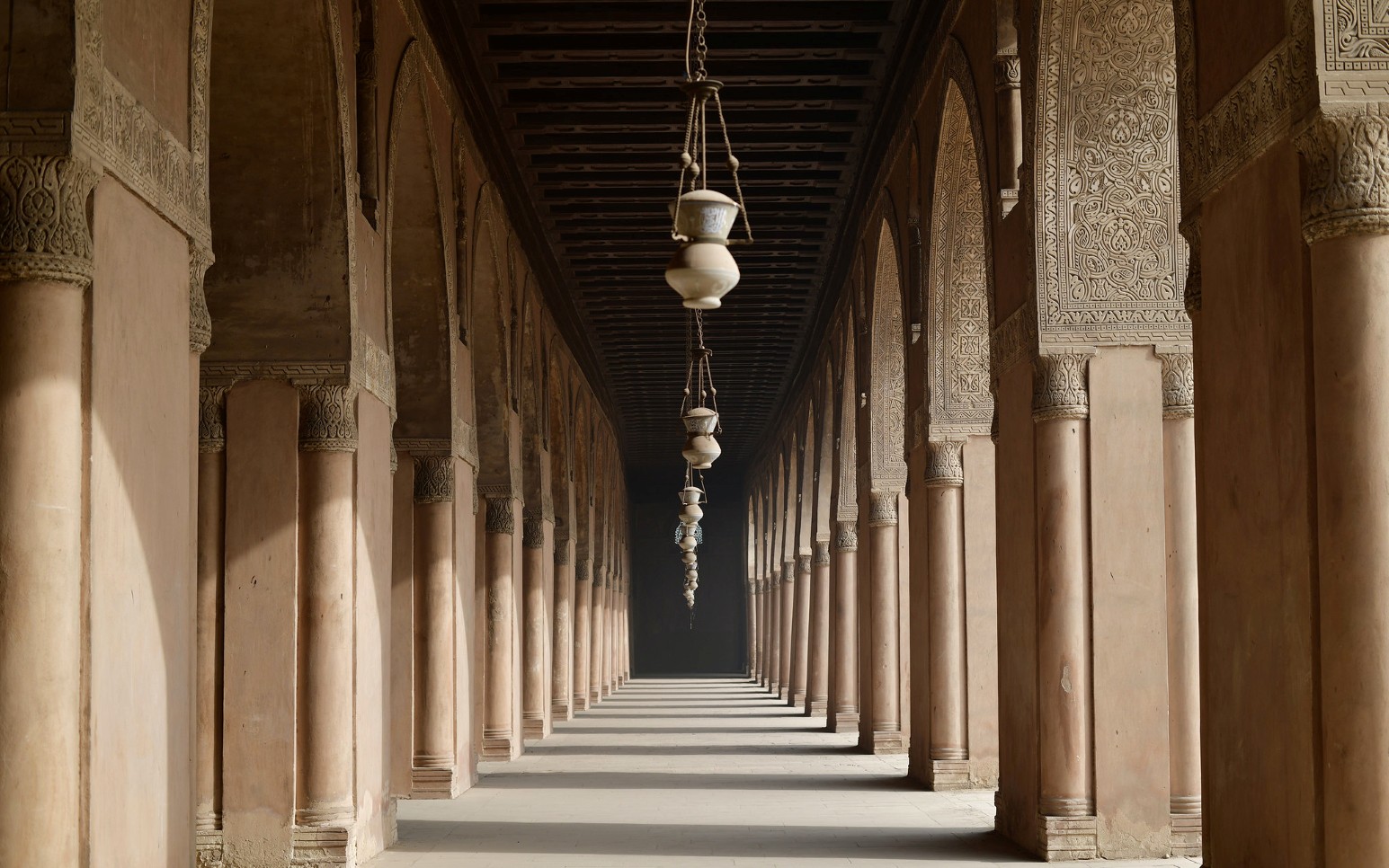 Long arched colonnade with carved stucco decoration and hanging lamps inside a historic mosque in Cairo, Egypt