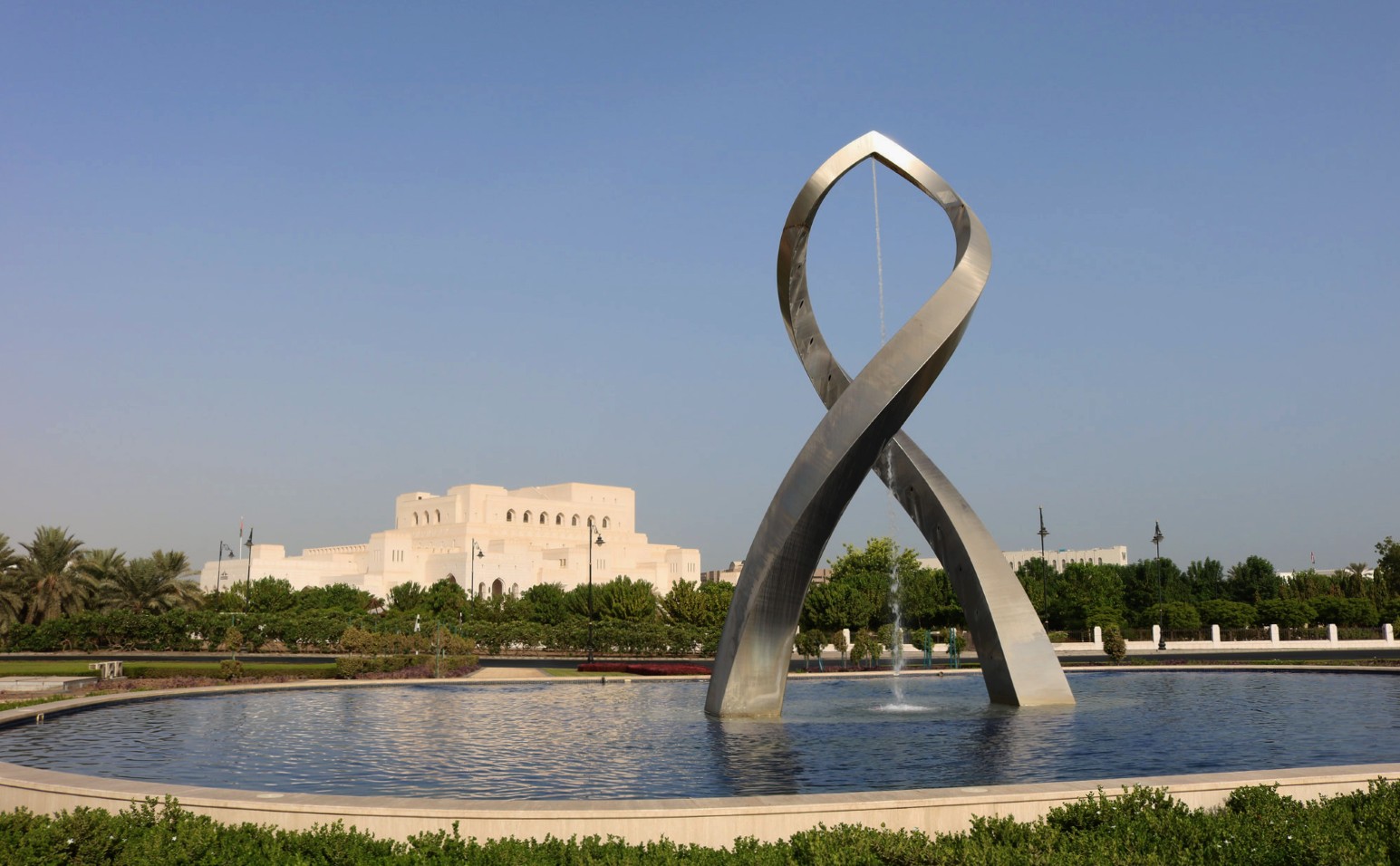 A twisting stainless steel sculpture rising from a circular fountain pool in Muscat, Oman, with the white Royal Opera House Muscat and palm trees behind