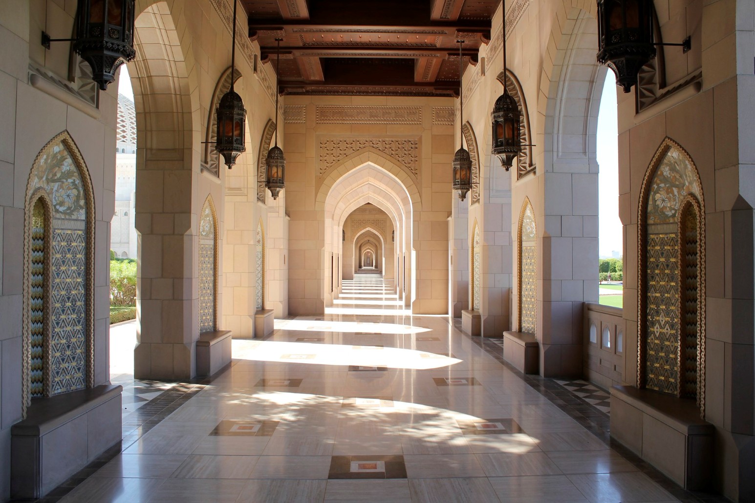 A long arcade of pointed stone arches, hanging lanterns and tiled wall panels at the Sultan Qaboos Grand Mosque in Muscat, Oman, with sunlight falling across the polished marble floor