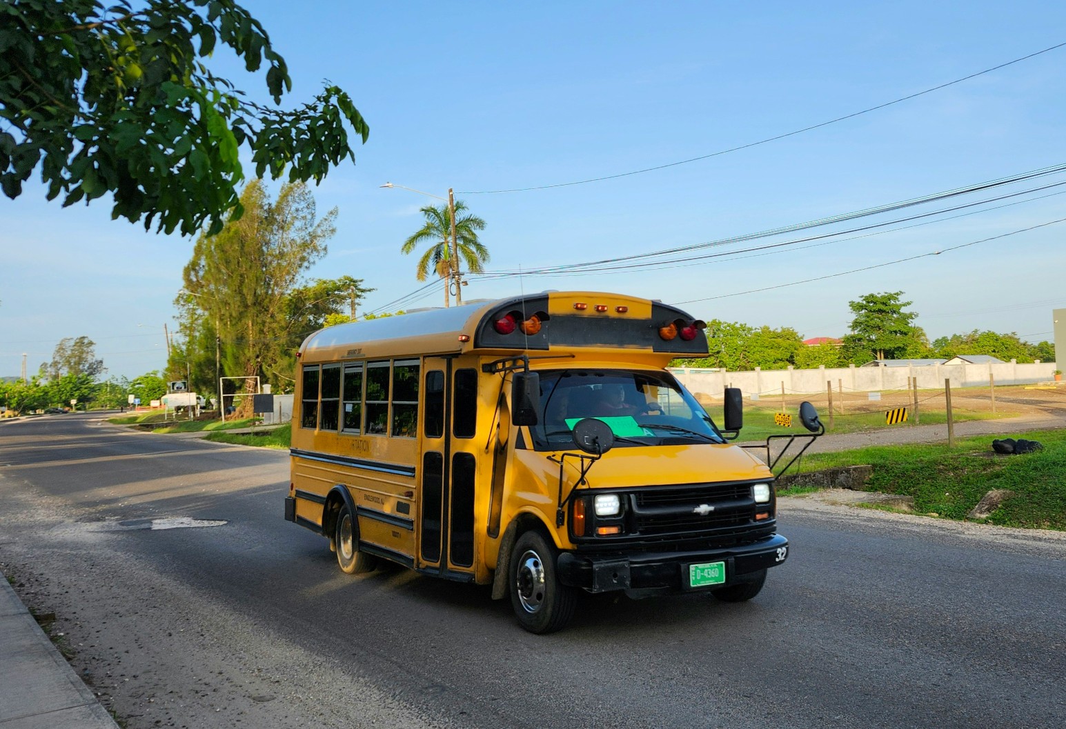A yellow bus driving along a quiet tree-lined road in Belmopan, Belize, under a clear blue sky with palm trees and power lines in the background